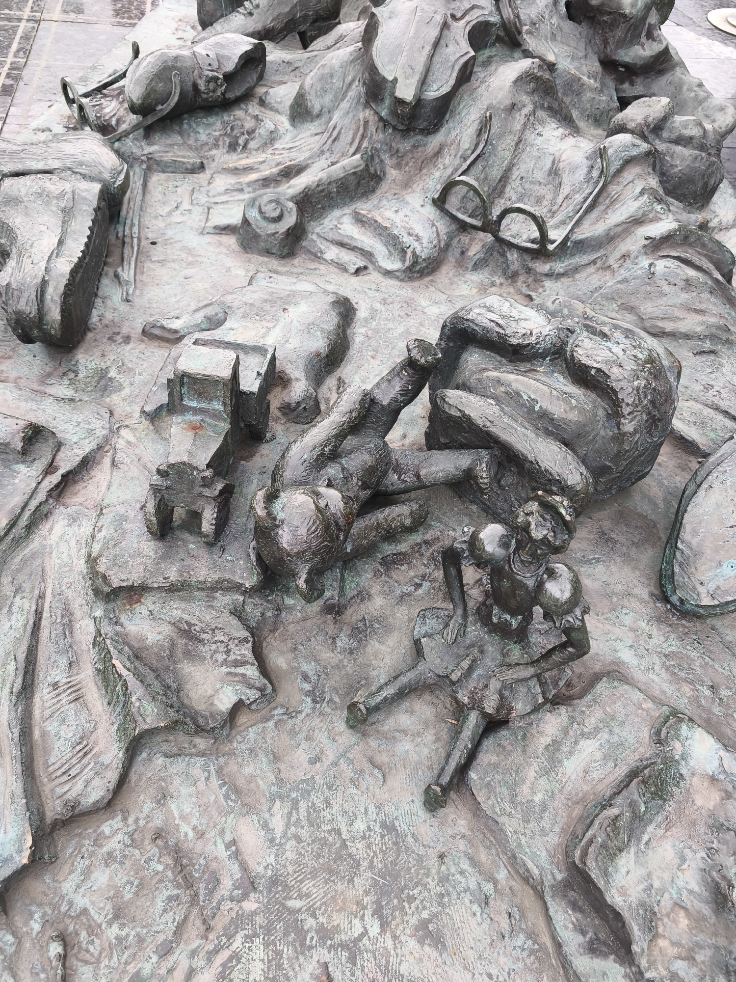 This image shows a close-up of a bronze bas-relief sculpture depicting several figures and objects. In the center, a stylized figure of a child in a dress and hat appears to be interacting with another figure lying on its back, with its legs outstretched. To the left of this scene, a small toy-like tractor is visible. Further to the left and upper left, there are other fragmented elements of figures and objects, including what appears to be a pair of glasses resting on a textured surface. The sculpture has a patinated bronze finish, with areas of verdigris suggesting age and exposure to the elements. The background appears to be cobblestone or paving stones. This sculpture is part of the "Children's Circle" memorial located in the Patriarch's Ponds district of Moscow, Russia.