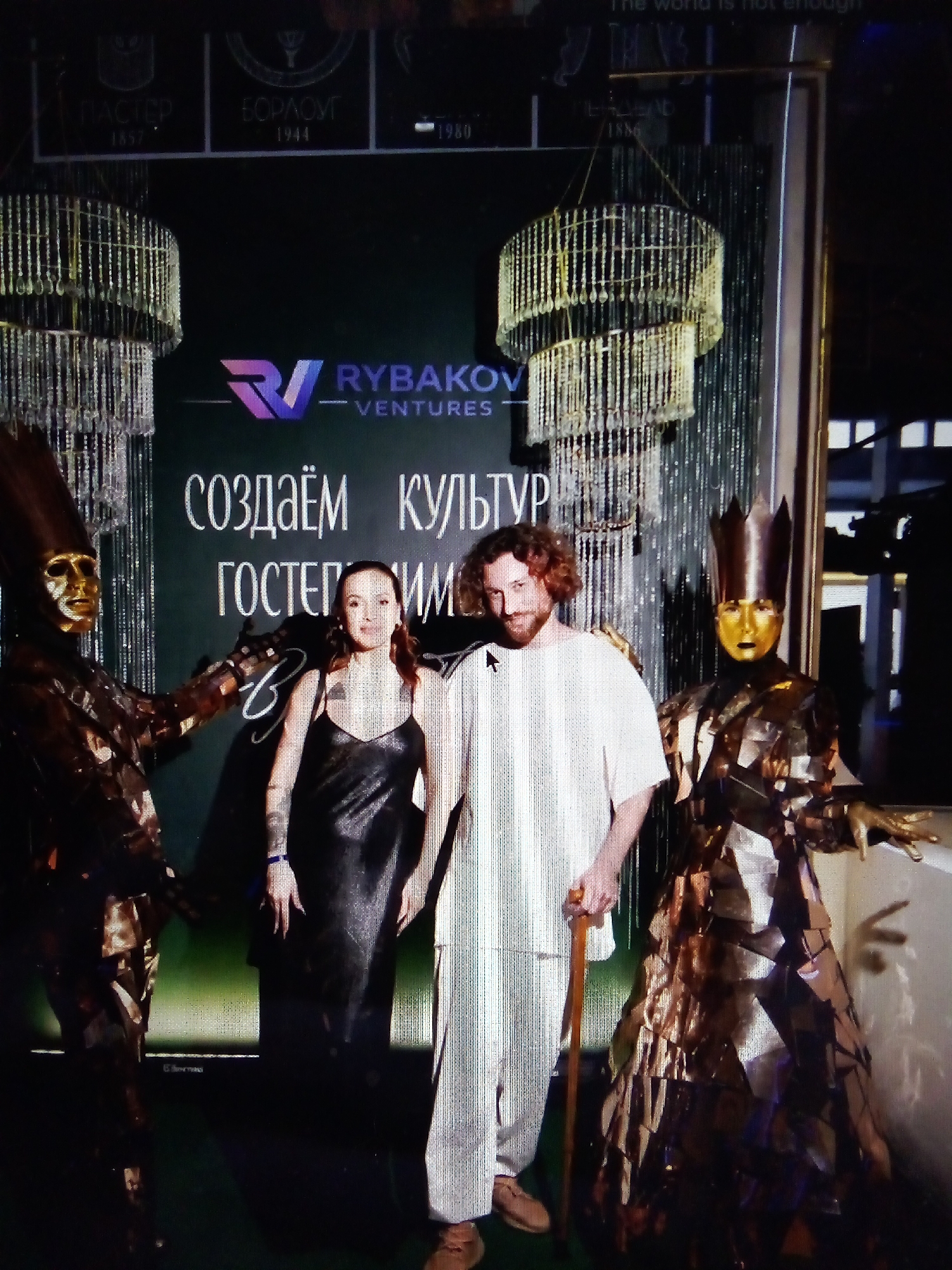 The image shows a red-haired man wearing a white t-shirt and pants, holding a cane, standing next to a woman in a black dress. They are flanked by two figures in elaborate, metallic costumes with golden masks. One costumed figure wears a crown. Behind them is a dark green backdrop with text in Russian, including "RYBAKOV VENTURES" and "СОЗДАЁМ КУЛЬТУРУ" (Creating Culture). Two crystal chandeliers hang from the ceiling. The event appears to be an outdoor or semi-outdoor gathering in a formal or artistic setting, possibly a red carpet event or a promotional display. The location is specified as посёлок дачного хозяйства «Архангельское» (Dacha Settlement "Arkhangelskoye"), Russia.