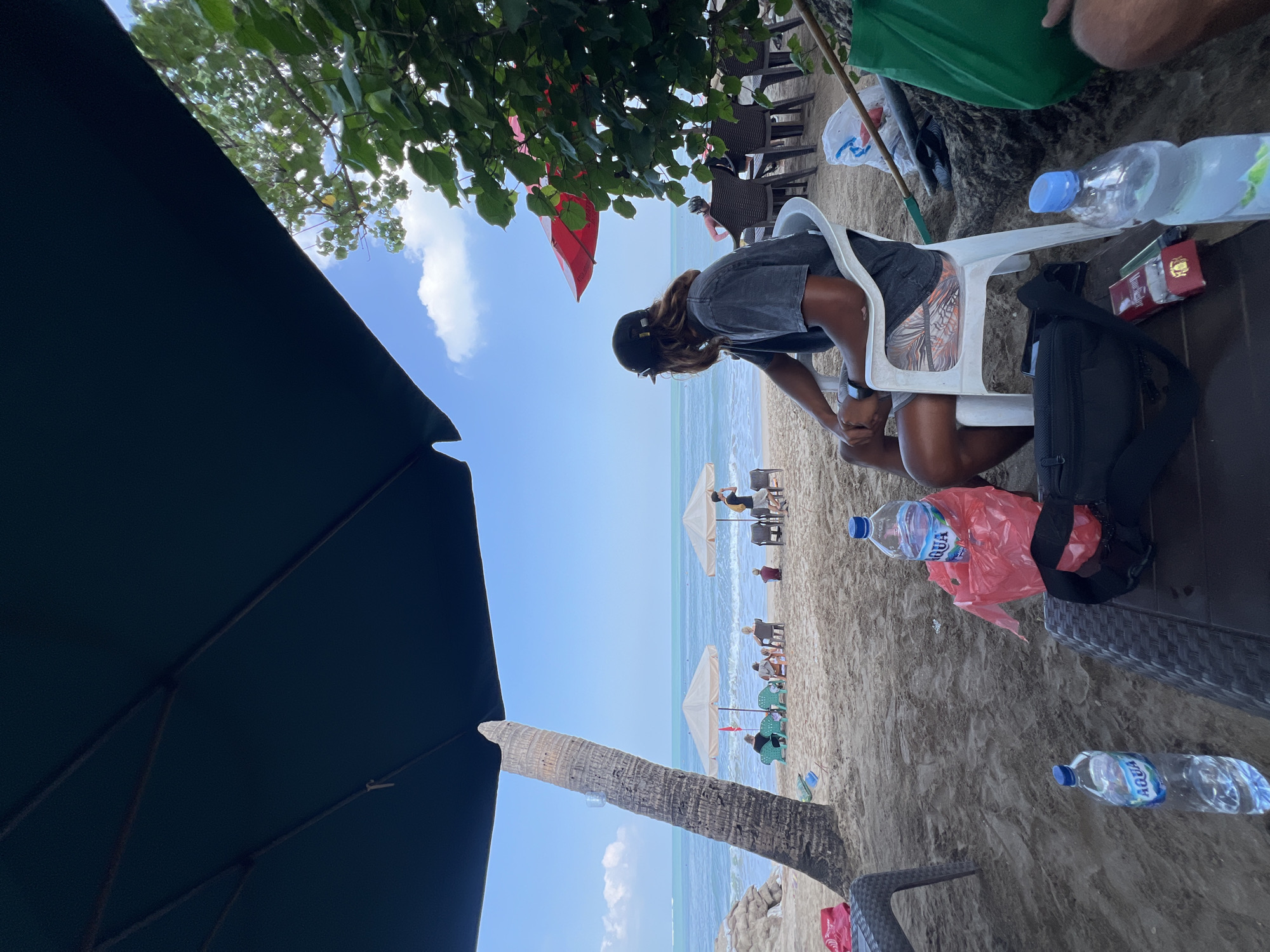 This is a medium shot, taken from under a large dark blue umbrella, of a beach scene in Kuta, Indonesia. The foreground shows a person sitting in a white plastic chair, facing away from the camera and looking out at the ocean. They are wearing a dark t-shirt and patterned shorts, and have long, light brown hair and a black baseball cap. To the left of the chair is a dark brown table with a black fanny pack and a pack of cigarettes on it. Two bottles of Aqua water are visible, one on the table and another near a red plastic bag. In the background, the sandy beach stretches towards the ocean. Several other people are seated on lounge chairs under umbrellas further down the beach. A palm tree stands on the left side of the frame. The ocean is calm with small waves, and the sky is blue with scattered white clouds. A red umbrella is visible in the background among green foliage. The scene appears to be a relaxed day at the beach.