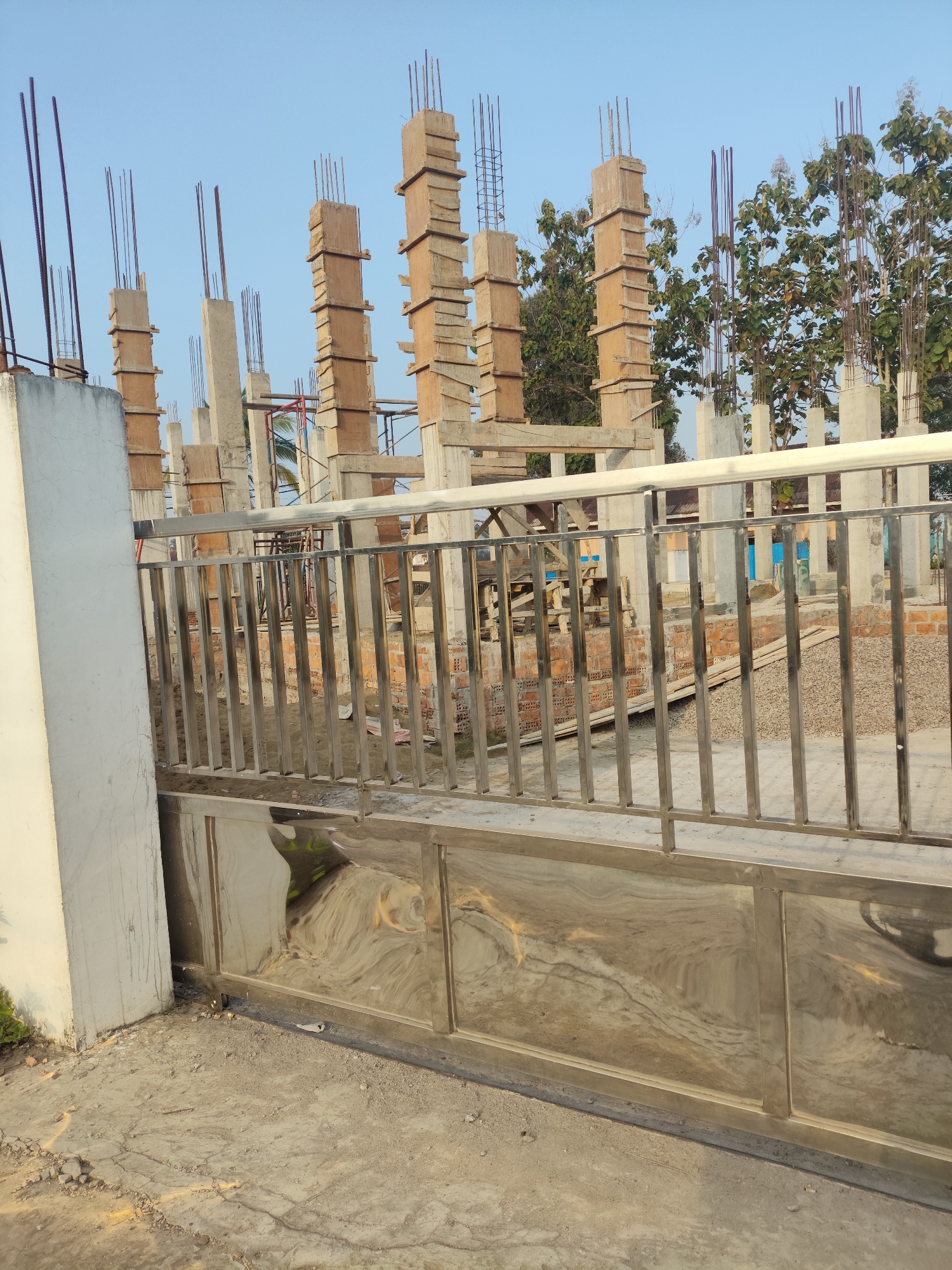This is a wide shot, taken from a low angle, of a construction site in Palembang, Indonesia. The foreground is dominated by a large, polished metal gate, likely for a residential or commercial property. Behind the gate, rows of concrete pillars with exposed rebar rise towards the sky, indicating ongoing construction of a multi-story building. Wooden formwork is still attached to some of the pillars, awaiting removal after the concrete has set. In the background, lush green trees suggest a tropical environment. The sky is a clear, pale blue, indicative of a sunny day. The ground is a mixture of concrete and loose gravel. The scene conveys a sense of development and progress.