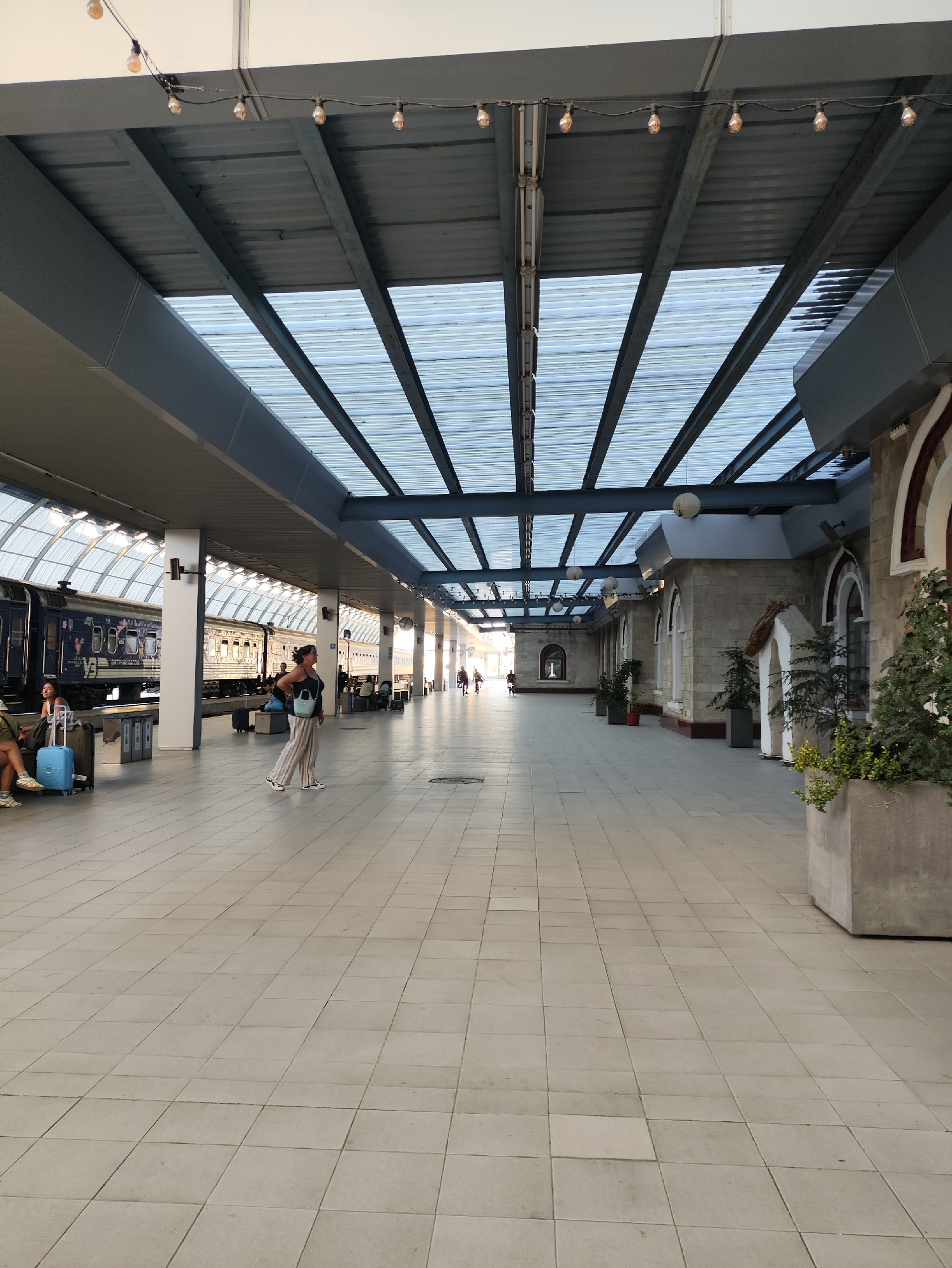 The image depicts a train station platform in Chișinău, Moldova. A train with a dark blue livery and "Airlines" written on its side is parked at the platform. Several people are visible, including a woman in a blue top and striped pants standing near the center of the image, a woman with short hair and a green jacket sitting on a bench with a blue suitcase, and other individuals in the background. The platform is covered by a translucent roof supported by a metal structure, with string lights illuminating the area. The floor is tiled with square beige tiles. Buildings with stone facades and arched windows are visible on the right side of the platform. Potted plants and large concrete planters are also present. The overall scene suggests a functional and somewhat modern train station environment.