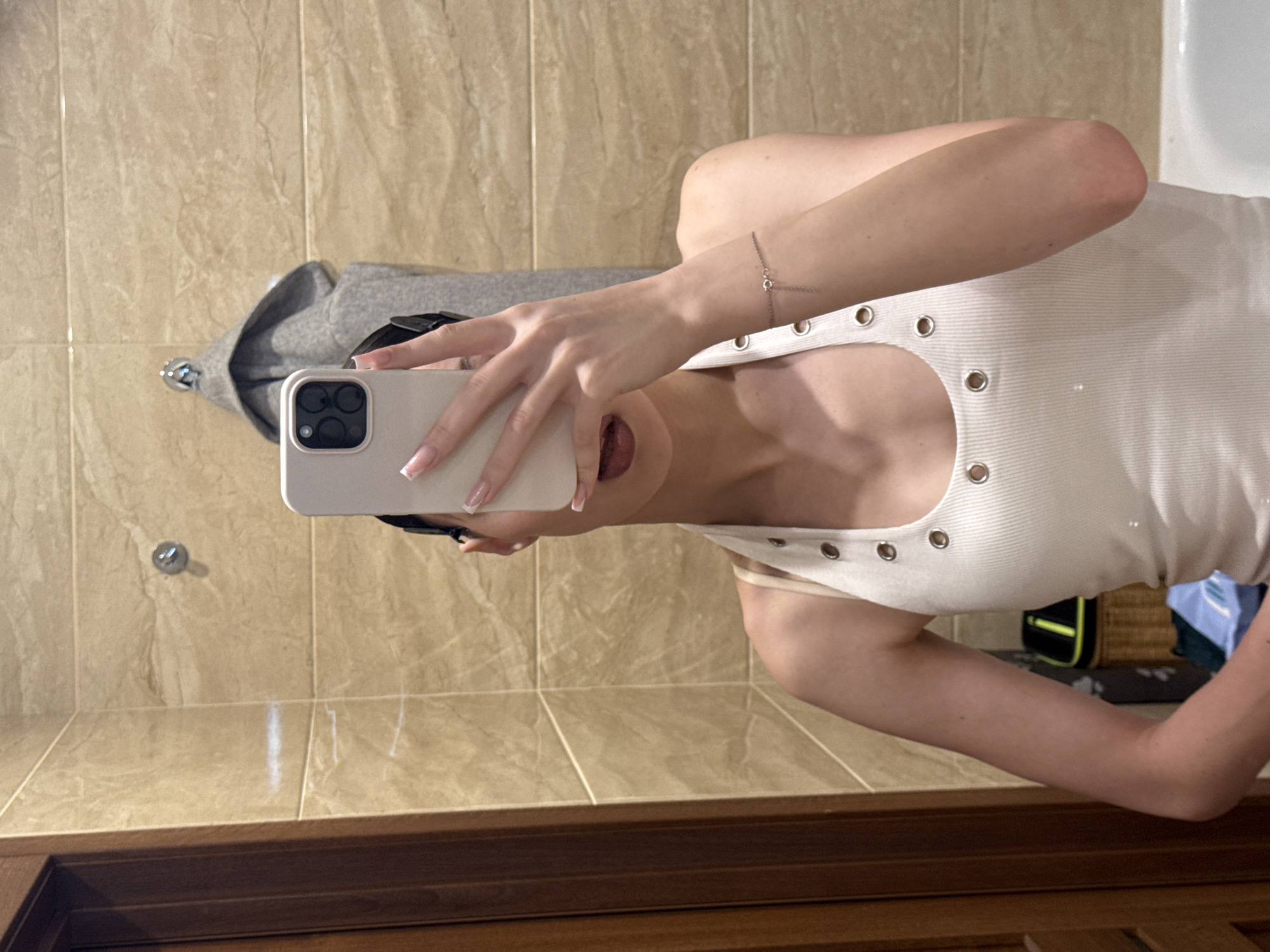A person is taking a selfie in a bathroom with beige tiled walls and a wooden cabinet below. The person is holding a white smartphone and is wearing a white, ribbed tank top with grommet details. They are also wearing a delicate silver bracelet on their left wrist. The person's right arm is bent and resting on their lap, and their left arm is extended to hold the phone. The background shows a reflection of the bathroom. The overall impression is a casual, private moment. No visible text is present in the image.