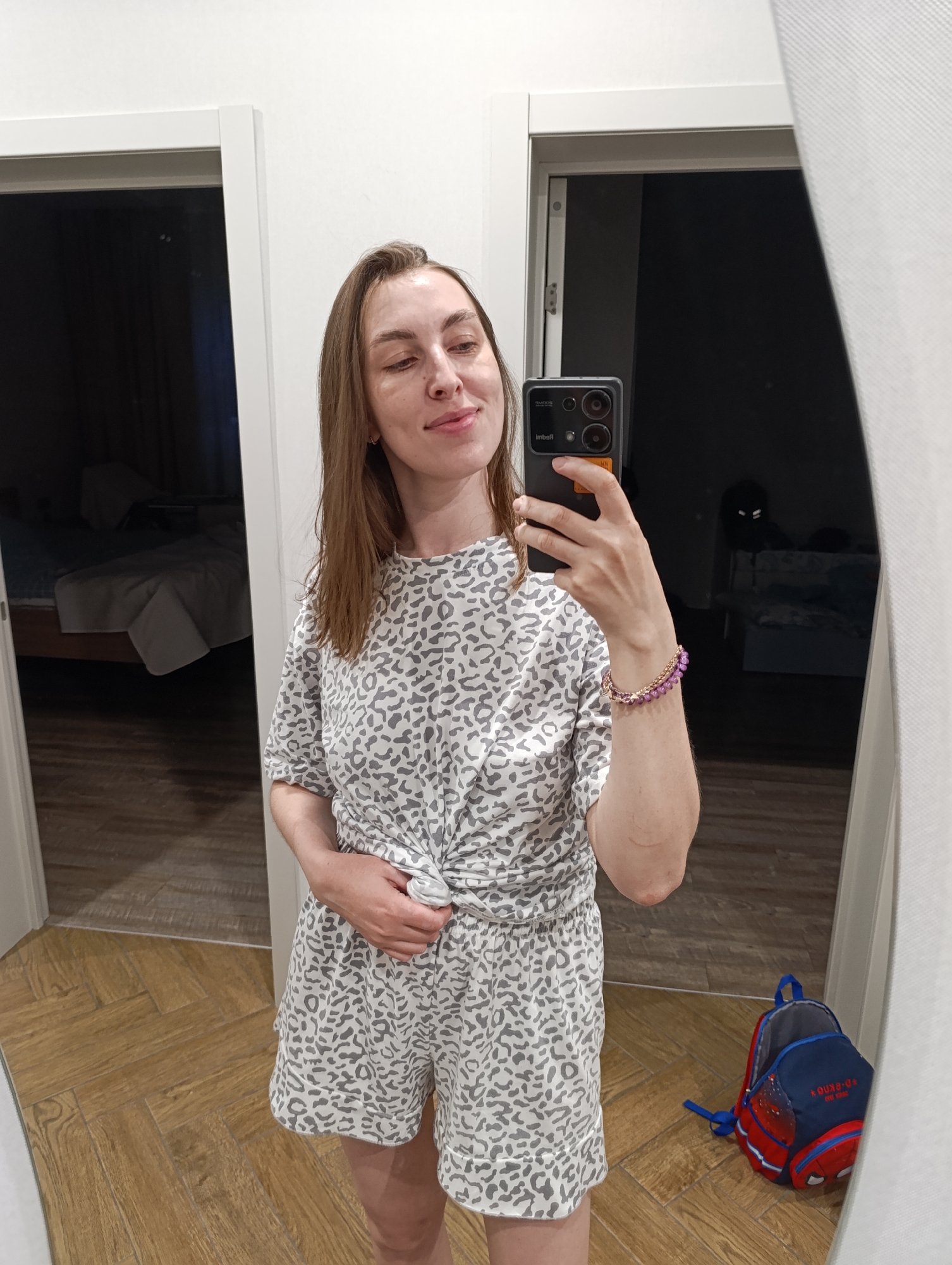 A woman is taking a selfie in a mirror. She is wearing a grey and white spotted pajama set and a purple bracelet. The setting is indoors, with two doorways visible. One doorway leads to a bedroom with a bed visible. There is a backpack on the floor. The location is Chaikovsky, Russia.