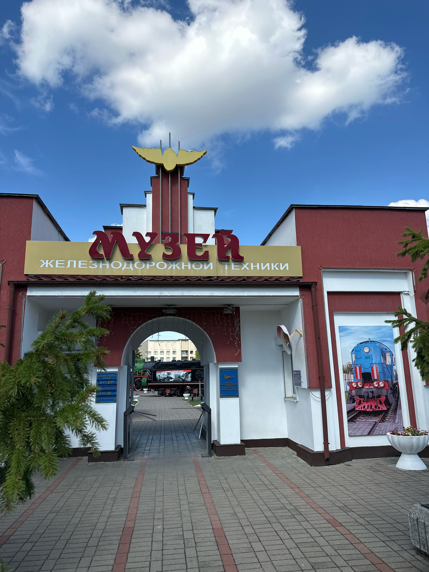 The image shows the entrance to the Brest Railway Museum in Brest, Belarus. The museum's sign is in Russian, displaying "МУЗЕЙ" (Museum) in large red letters and "ЖЕЛЕЗНОДОРОЖНОЙ ТЕХНИКИ" (Railway Equipment) in smaller white letters below. The building features a stylized golden bird sculpture on its facade. Through an arched entrance, vintage trains are visible on display. To the right of the entrance, a large mural depicts a blue and red locomotive. The ground in front of the museum is paved with brickwork, and a pine tree stands to the left. The sky above is blue with scattered white clouds.