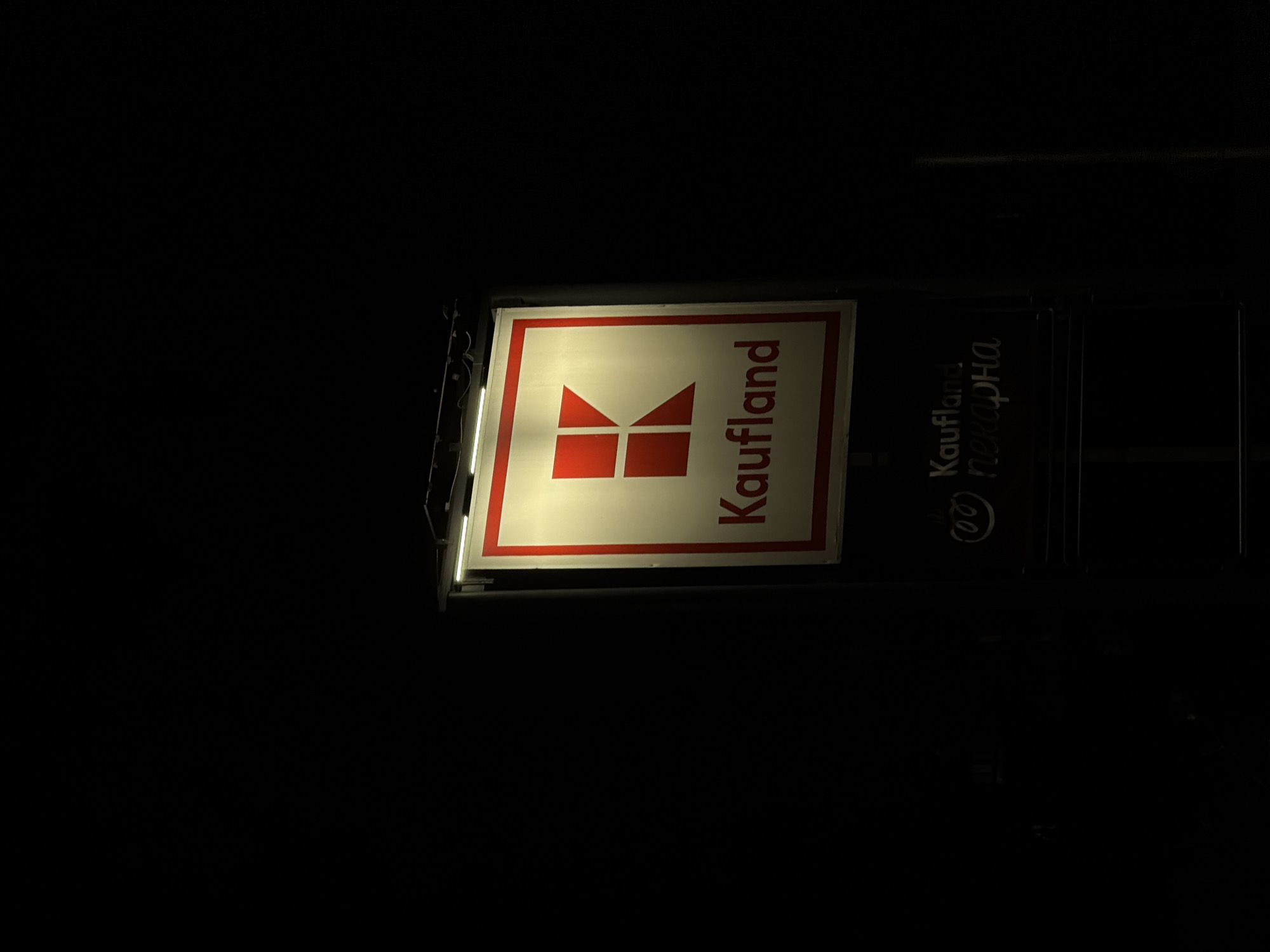 The image displays a brightly illuminated "Kaufland" supermarket sign at night in Sofia, Bulgaria. The sign is primarily composed of two sections: a prominent, brightly lit white square with a bold red border, and a darker, less illuminated section to its right.
On the main illuminated part of the sign, the distinctive red Kaufland logo is centered, which appears as a stylized 'K' or four interlocking red shapes. To the right of the logo, the brand name "Kaufland" is written vertically in red, sans-serif letters.
On the darker section to the right, some text is visible, including "Kaufland" in Cyrillic script, followed by the Cyrillic word "пекарна" (bakery). This text is lighter against the dark background but not as brightly lit as the main sign. The surrounding environment is almost entirely black, emphasizing the artificial light of the sign against the darkness of the night. Illumination strips are visible on the left side of the main sign, providing light. No people or activities are visible, and the focus is solely on the illuminated commercial signage.