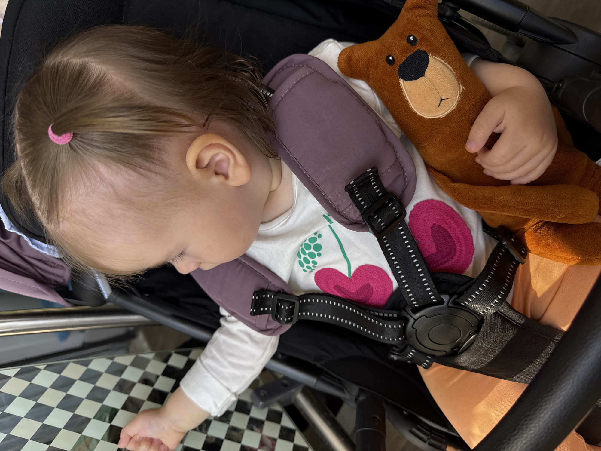 A toddler lies asleep in a stroller, secured with a purple-padded harness, clutching a brown stuffed bear with a tan muzzle. The child has fair skin, light brown hair held back with a pink hair clip, and is wearing a white shirt with pink floral or cherry blossom designs. The stroller has a black frame and a black and white checkered footrest. The location is not identifiable.