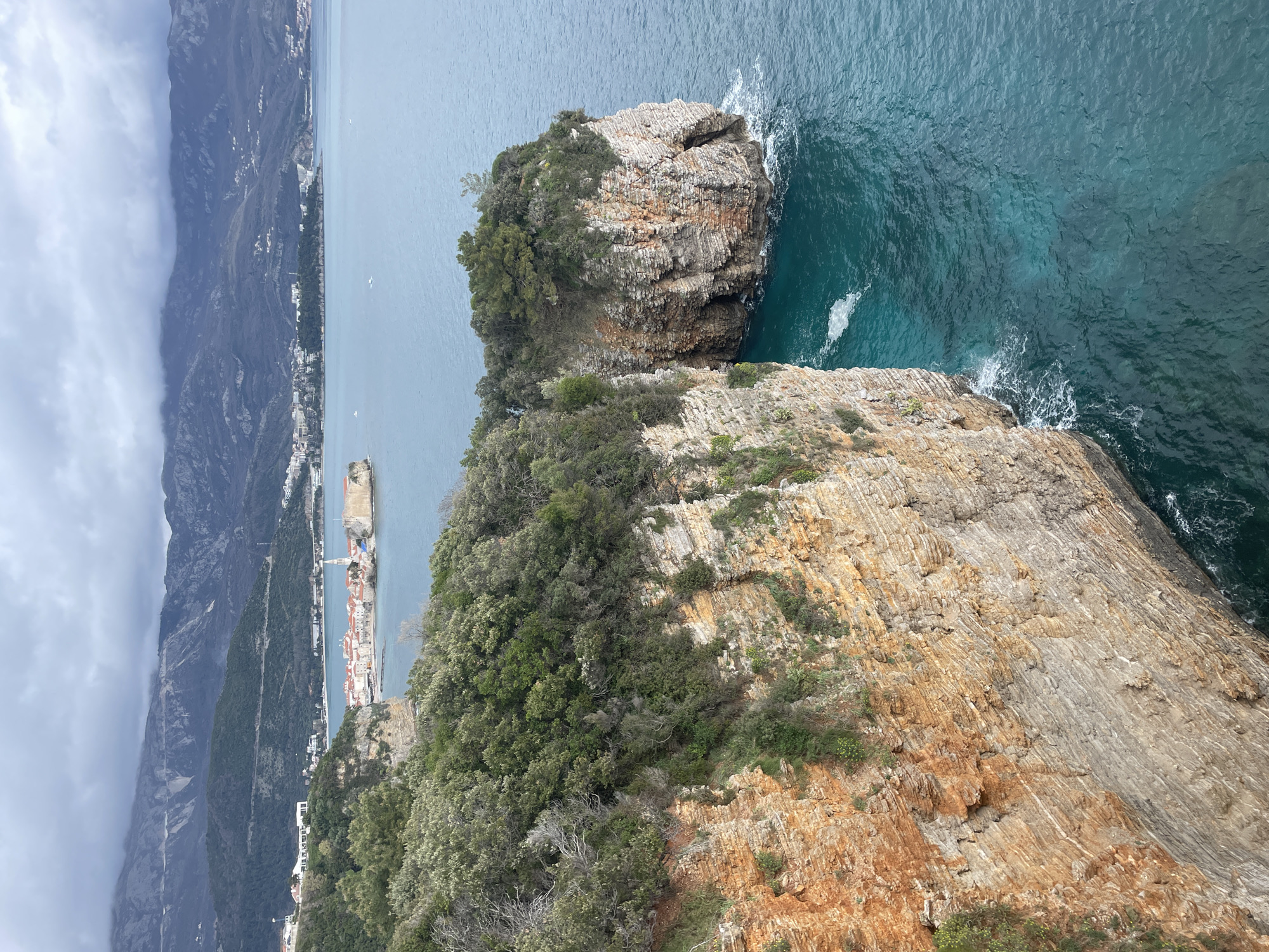 The image presents an elevated view of the coastline near Budva, Montenegro. In the immediate foreground to the left, a sheer cliff face composed of stratified orange and grey-brown rock drops vertically into the turquoise-blue sea. The cliff top and ledges are covered in dense green Mediterranean vegetation. To the right, a separate, smaller rocky outcrop, similarly vegetated, emerges from the water, with white foam indicating wave activity at its base.
The midground features the calm, deep blue-green waters of the Adriatic Sea. In the distance, prominently situated on a small peninsula, is the Old Town of Budva, characterized by its cluster of red-tiled roofs, white stone buildings, and a tall bell tower. Modern buildings line the coast on either side of the Old Town peninsula.
The background consists of a chain of dark, rugged mountains that rise steeply behind the coastal settlements. The upper slopes and peaks of these mountains are partially shrouded by a heavy, overcast sky filled with grey clouds. No individuals or specific human activities are visible within the frame.