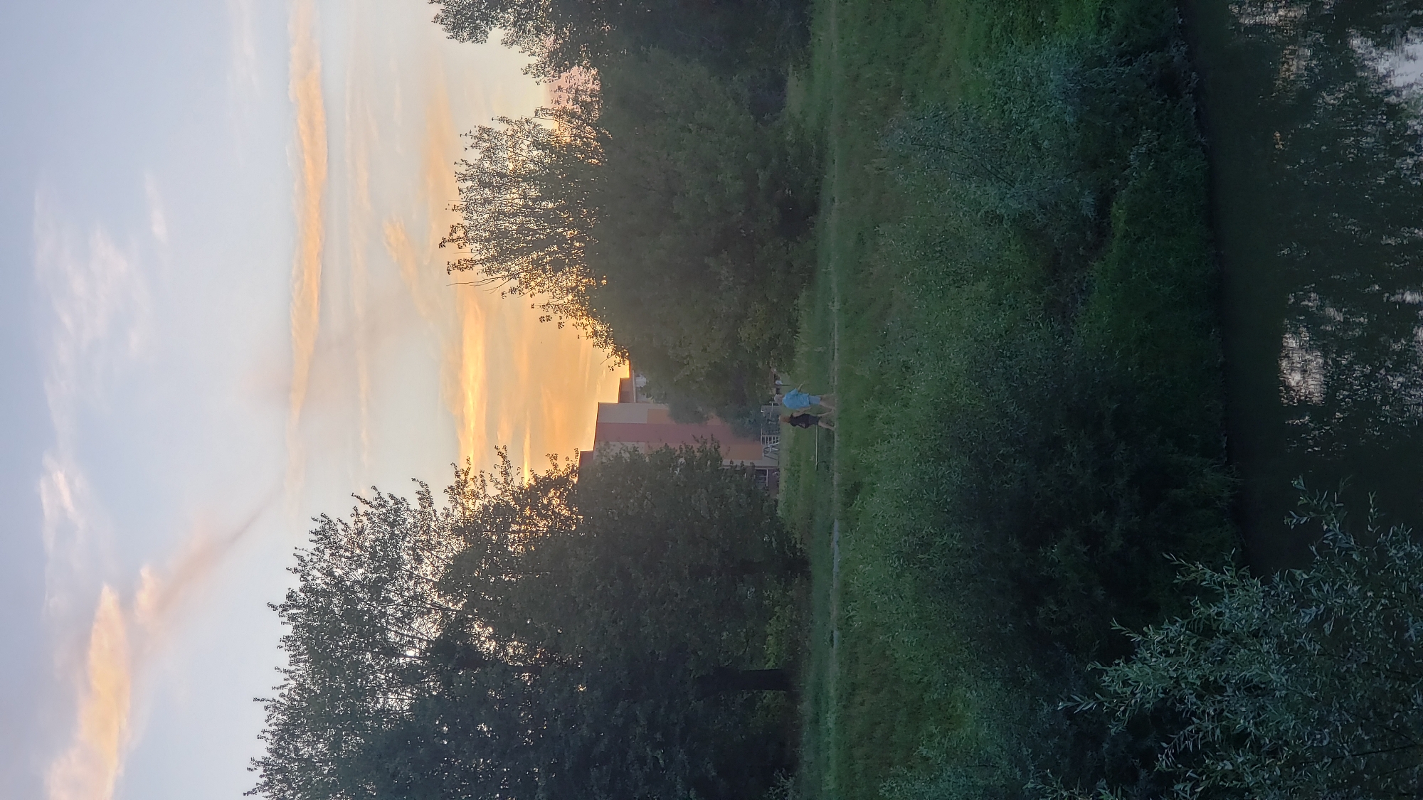 The image depicts a sunset scene in Prešov, Slovakia, likely near the Sekčov riverbank. In the foreground, a dark body of water reflects the sky and surrounding vegetation. The middle ground features a grassy area with dense green bushes and trees. Two people are walking on a path, one in a dark dress and the other in a light blue shirt and shorts. Behind them, a multi-story building with orange and pink facades is visible, partially obscured by trees. The sky is a pale blue with wispy clouds tinged with orange and yellow hues from the setting sun. A notable cloud formation is a thin, dark streak extending from the upper left quadrant of the sky.