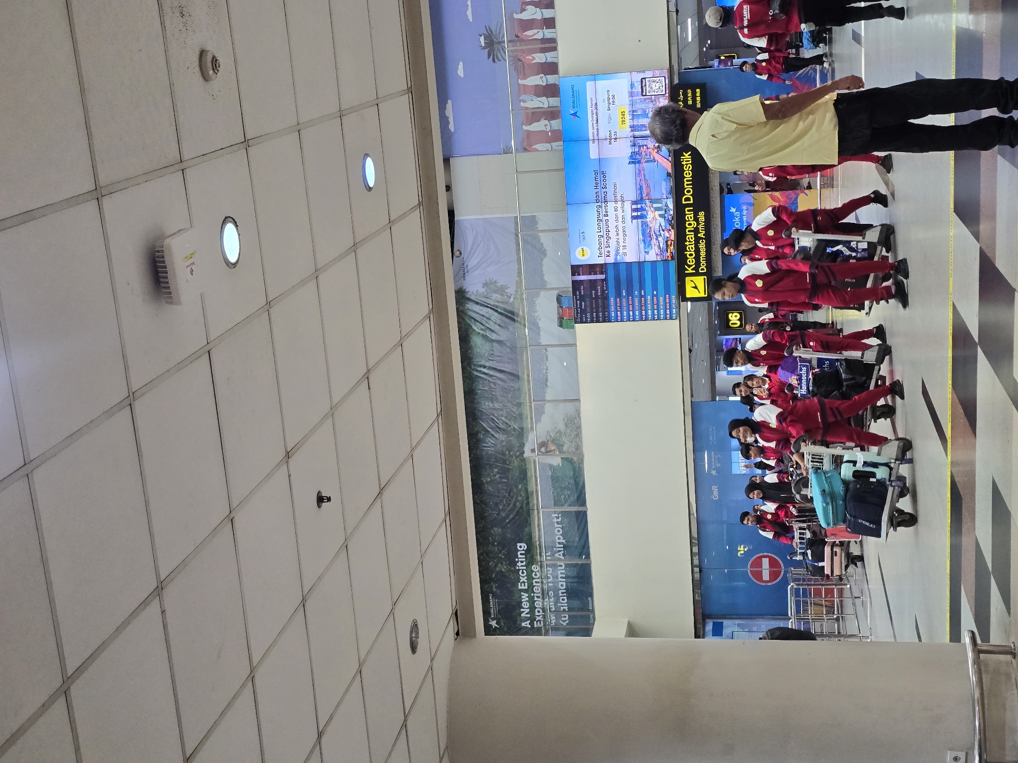 The image depicts the interior of an airport terminal, specifically within the domestic arrivals area. A group of individuals, predominantly young adults, are seen wearing matching red and white athletic uniforms, suggesting they are a sports team or delegation. They are pulling luggage carts laden with bags, indicating they are arriving or departing.
In the background, a large digital display board shows flight information and advertisements. One prominent advertisement displays "Kualanamu Airport: A New Exciting Experience". Another section of the board advertises "Terbang Langsung dan Hemat! Ke Singapura Bersama Scoot!", which translates to "Fly Direct and Save! To Singapore with Scoot!". The overall environment is brightly lit, with a tiled ceiling and floor, characteristic of a modern airport. The atmosphere appears busy, with other travelers and airport personnel visible in the periphery. The presence of airport signage like "Kedatangan Domestik Domestic Arrivals" confirms the location as an airport's arrival hall. The overall scene captures a moment of transit, with the group of uniformed individuals seemingly processing their arrival or preparing for departure.