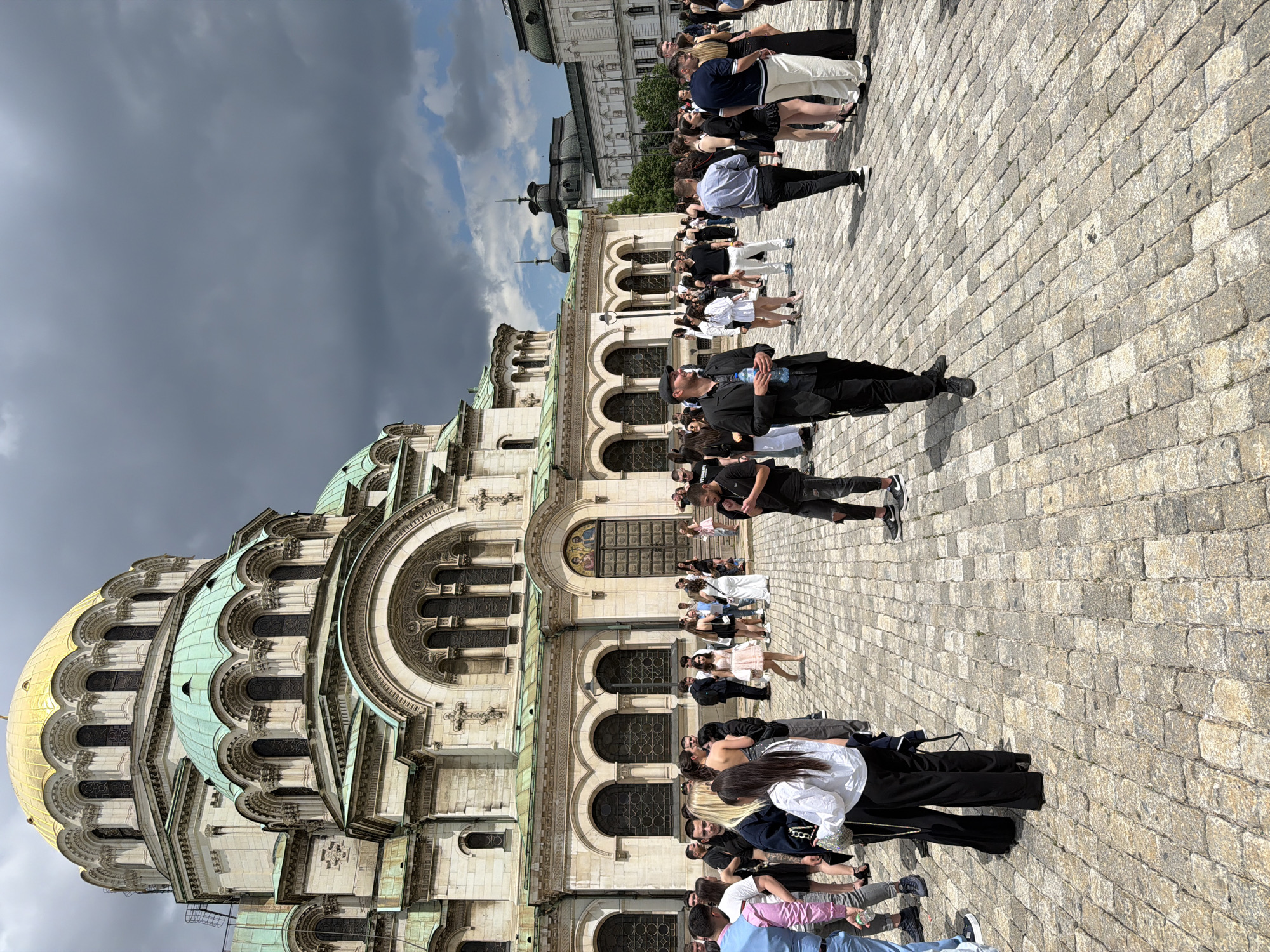 The image depicts a crowd of people outside the Alexander Nevsky Cathedral in Sofia, Bulgaria. The cathedral's gold dome and green roof are prominently visible. People stand on a paved square, some appear to be tourists, and there are architectural details of the building in the background. The sky above shows overcast conditions.