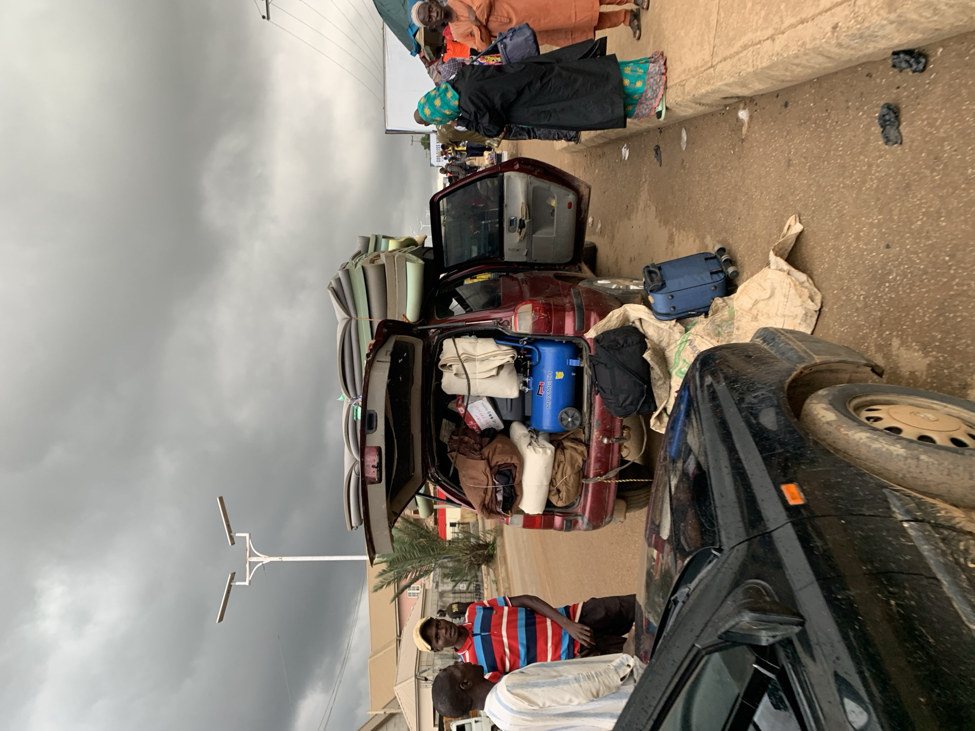 The image depicts an outdoor scene in the city of Rafin Par, Nigeria, on a cloudy day. The focal point is a maroon minivan with its rear hatch and driver's side door open, heavily laden with luggage and personal belongings. On the roof of the minivan, several large foam mattresses are stacked. Inside the open rear hatch, various bags, rolled blankets, and a blue air compressor are visible. A black car is parked in the foreground, partially obscuring the view of the minivan's front.
To the left of the minivan, two men stand conversing. One, wearing a white and blue striped shirt and a yellow cap, is facing the viewer. The other, wearing a white striped garment, is facing the first man.
To the right of the minivan, a woman in a black abaya and a green headscarf is standing, facing away from the viewer. Further to the right, a man in an orange garment and cap stands with his arms crossed, also facing away from the viewer. Several other individuals are visible in the background, suggesting a bustling area.
The ground is a dirt road, and a concrete curb is visible on the right side of the image. Bags and a blue suitcase are scattered on the ground near the minivan. The sky is overcast with gray clouds.