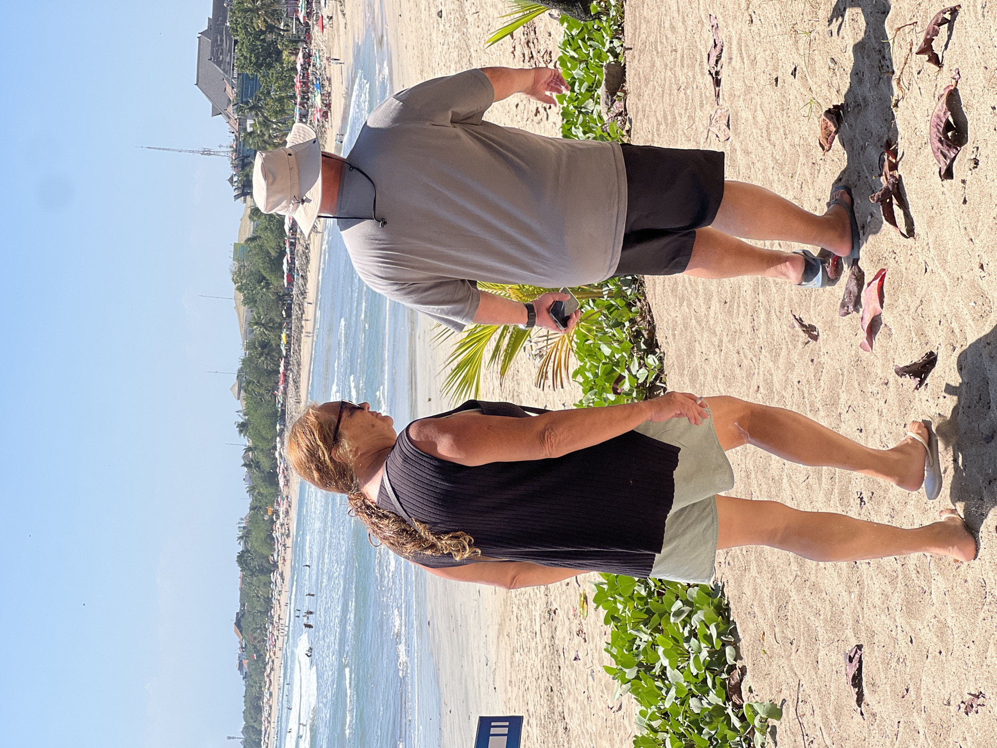 Two people, a woman and a man, walk on the beach in Kuta, Indonesia. The woman, with long auburn hair styled in a braid, wears a black sleeveless top and light green shorts. She has sunglasses on and is wearing white flip-flops. The man, wearing a light gray t-shirt and black shorts, has a light-colored bucket hat on. He is wearing sandals and holds a smartphone in his right hand. They are walking away from the camera towards the ocean, which is visible in the background. The beach is sandy and lined with lush green vegetation in the foreground. The ocean has small waves, and several people are visible in the water and on the beach, with many colorful umbrellas dotting the sand. Buildings and palm trees are visible in the distance along the coastline. The sky is clear and blue.
