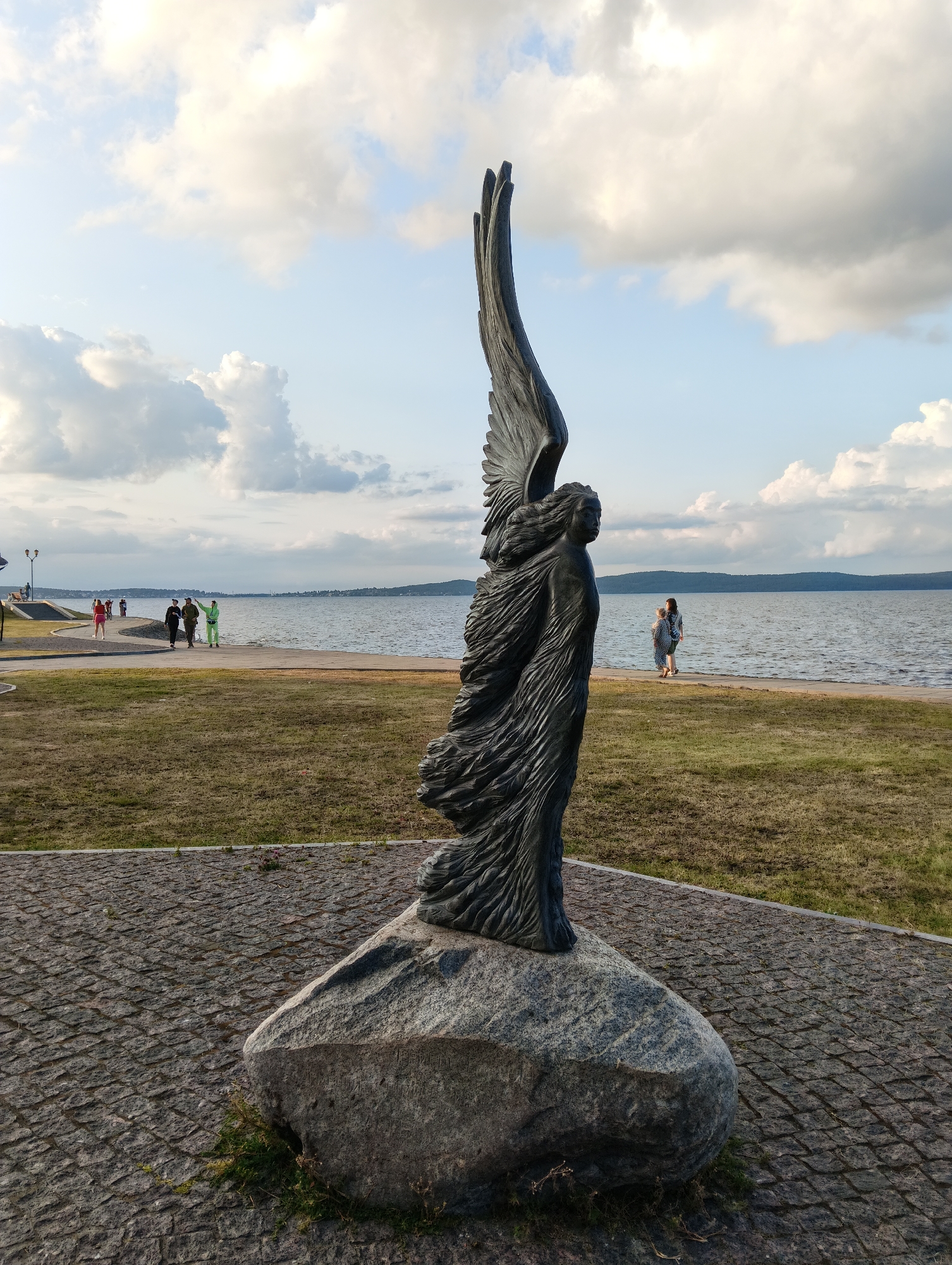 A dark, weathered bronze statue of a winged figure stands on a large, rough-hewn grey boulder. The figure's wings are spread upwards, and its body appears to be in motion, with long, flowing drapery or feathers. The statue is positioned on a cobblestone plaza. In the background, a wide body of water, likely Lake Onega, stretches to the horizon. Along the shoreline, a paved pathway is visible, with several individuals walking. The pathway is bordered by a strip of dry grass, which then transitions into a larger grassy area. The sky above is a mix of blue and large, white, fluffy clouds, indicating daytime. In the distance, a hazy shoreline with hills is discernible. The overall scene is outdoors and appears to be a public park or waterfront area in Petrozavodsk, Russia, possibly the Onega Embankment, given the presence of Lake Onega and the style of the statue, which is reminiscent of the "Dream" sculpture located there.