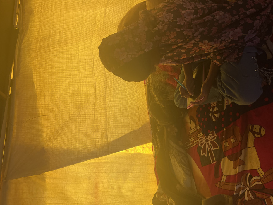 The image shows a young woman sitting inside a tent, writing in a notebook. The tent is made of yellow canvas. The woman is wearing a long floral-patterned garment. The canvas is stretched taut and has a grid-like texture. The woman is sitting on a red and black patterned blanket. The woman’s back is to the camera. She is holding a blue pen in her hand. The coordinates provided correspond to a location in Sahibzada Ajit Singh Nagar, India. The location of this photograph cannot be determined definitively based on the image alone. The image is shot in a low-light environment, with the light source outside the tent. The image appears to have been taken with a cell phone camera. The image appears to be in focus. The photo was taken in the afternoon, as it is backlit. The image is slightly blurry due to the motion of the person.
