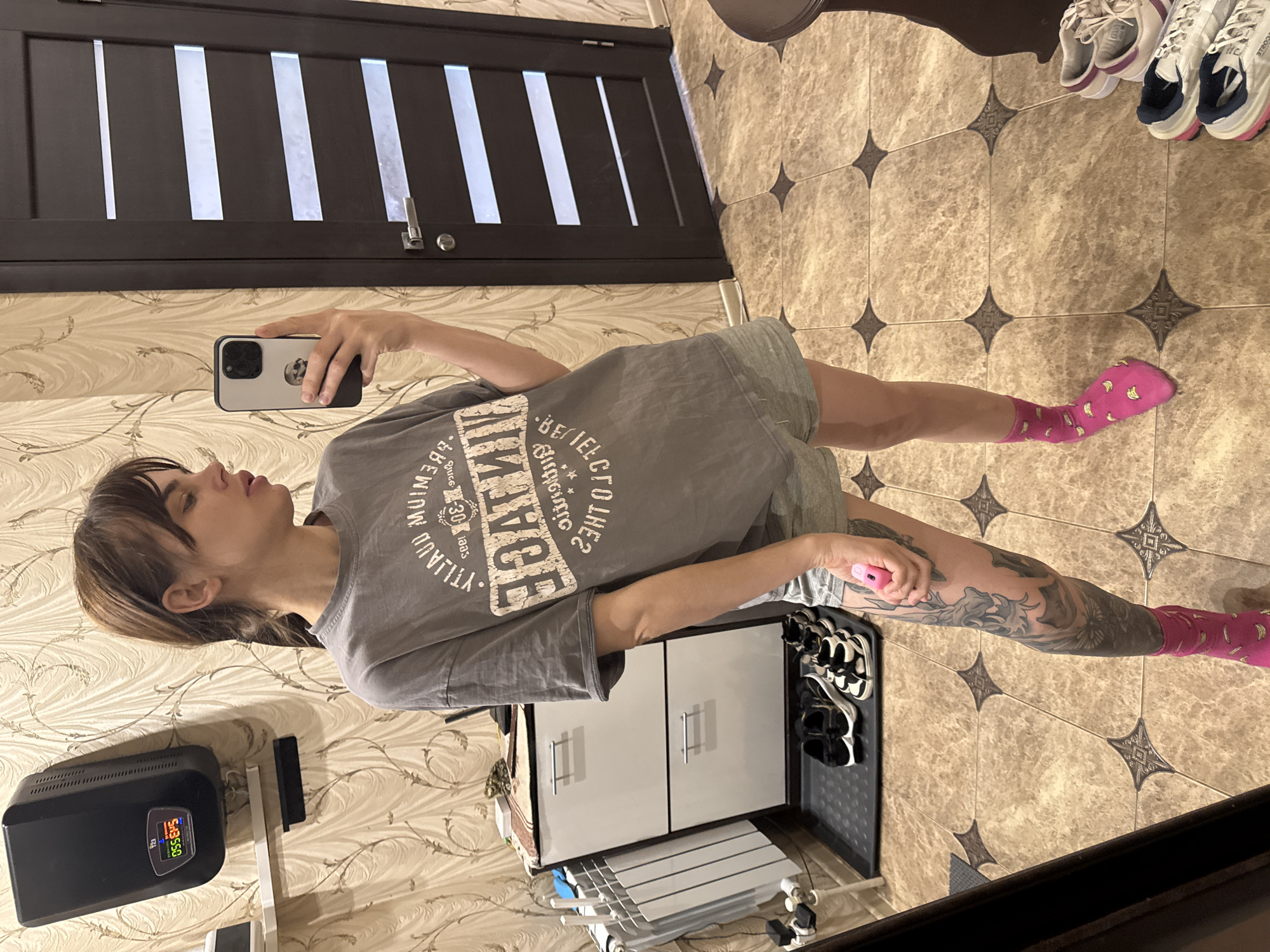 A person is taking a selfie in what appears to be a bathroom or entryway. The person is wearing a gray t-shirt, shorts, and pink socks with a banana pattern. They are holding a smartphone in their left hand and holding a pink object in their right. A portion of their tattooed leg is visible.
Behind the person is a white shoe cabinet with a black tray holding multiple pairs of shoes and sandals. Above the shoe cabinet is a clothing rack or drying rack.
To the left is a wall with a patterned wallpaper, featuring a black surge protector and a shelf. In the background, there is a dark brown door with horizontal white slats. The floor is tiled with a beige patterned design.
There is no indication of Rostov-on-Don, Russia in the image.