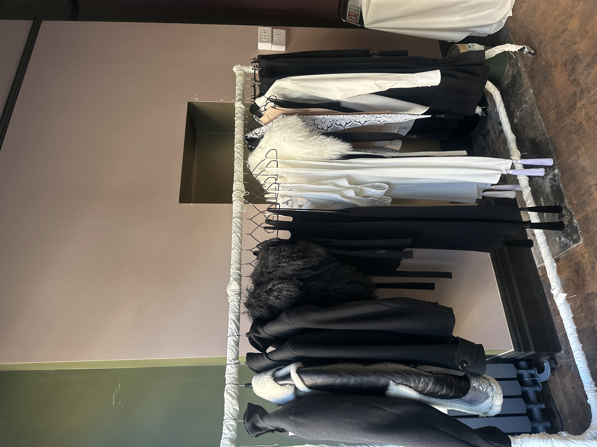 The image shows a rack of black and white clothing items, predominantly dresses and coats, displayed on hangers in what appears to be a studio or retail space. The backdrop consists of a pale mauve wall with a dark rectangular opening. To the left, a section of the wall is painted olive green. The clothing items vary in texture and style, with some featuring shearling or faux fur accents, and others appearing to be made of delicate lace or silk. The floor is made of wooden planks. There are no people visible in the image. The scene is clean and organized, suggesting a professional setup for displaying fashion items. Without further context, the specific location in London, United Kingdom, cannot be determined from the visual information alone.