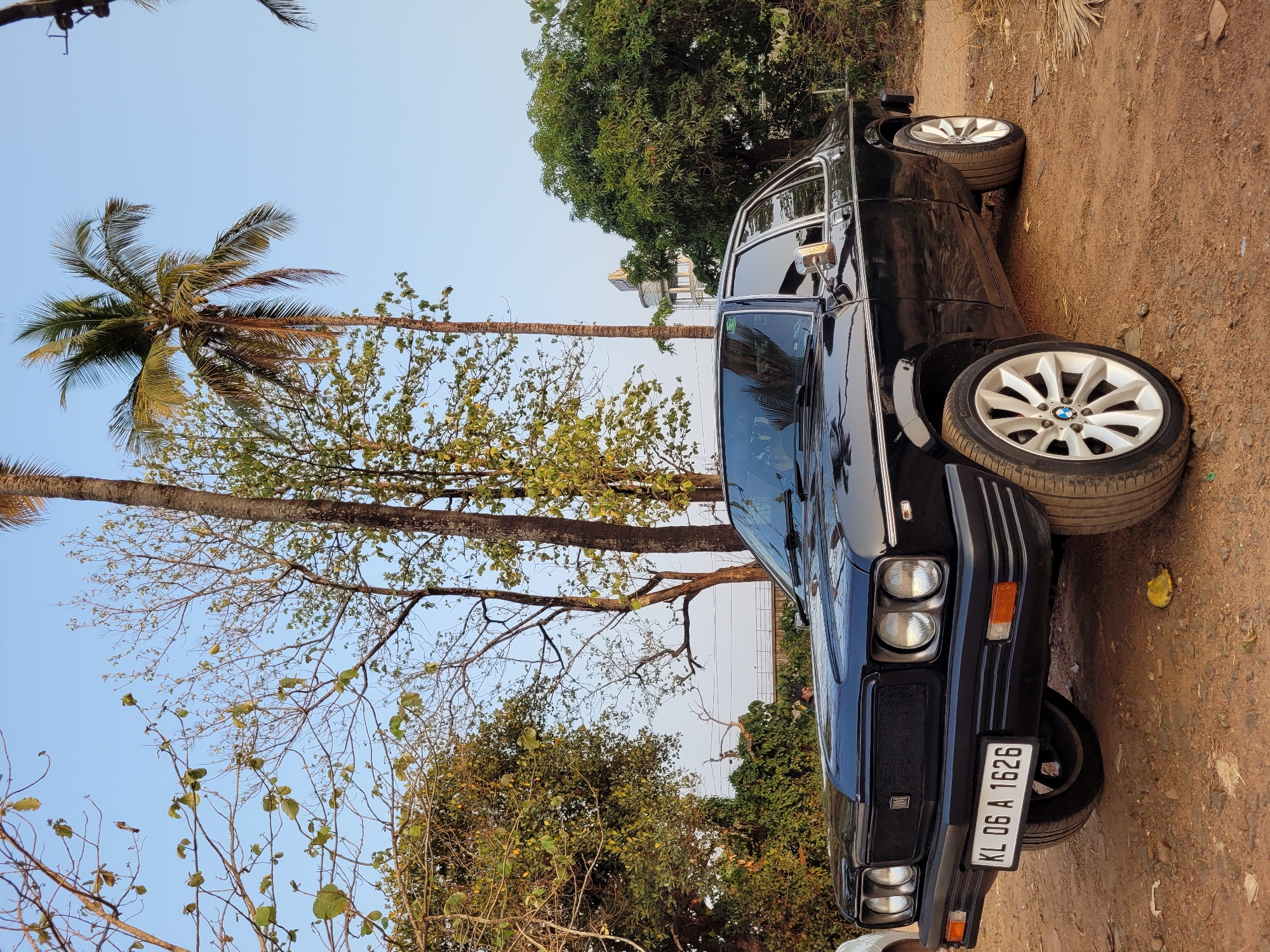 The image shows a black car parked on a dirt road. A palm tree is visible on the left side of the frame. The car has a license plate that reads "KL 06 A 1626." The location is Unknown, Unknown.