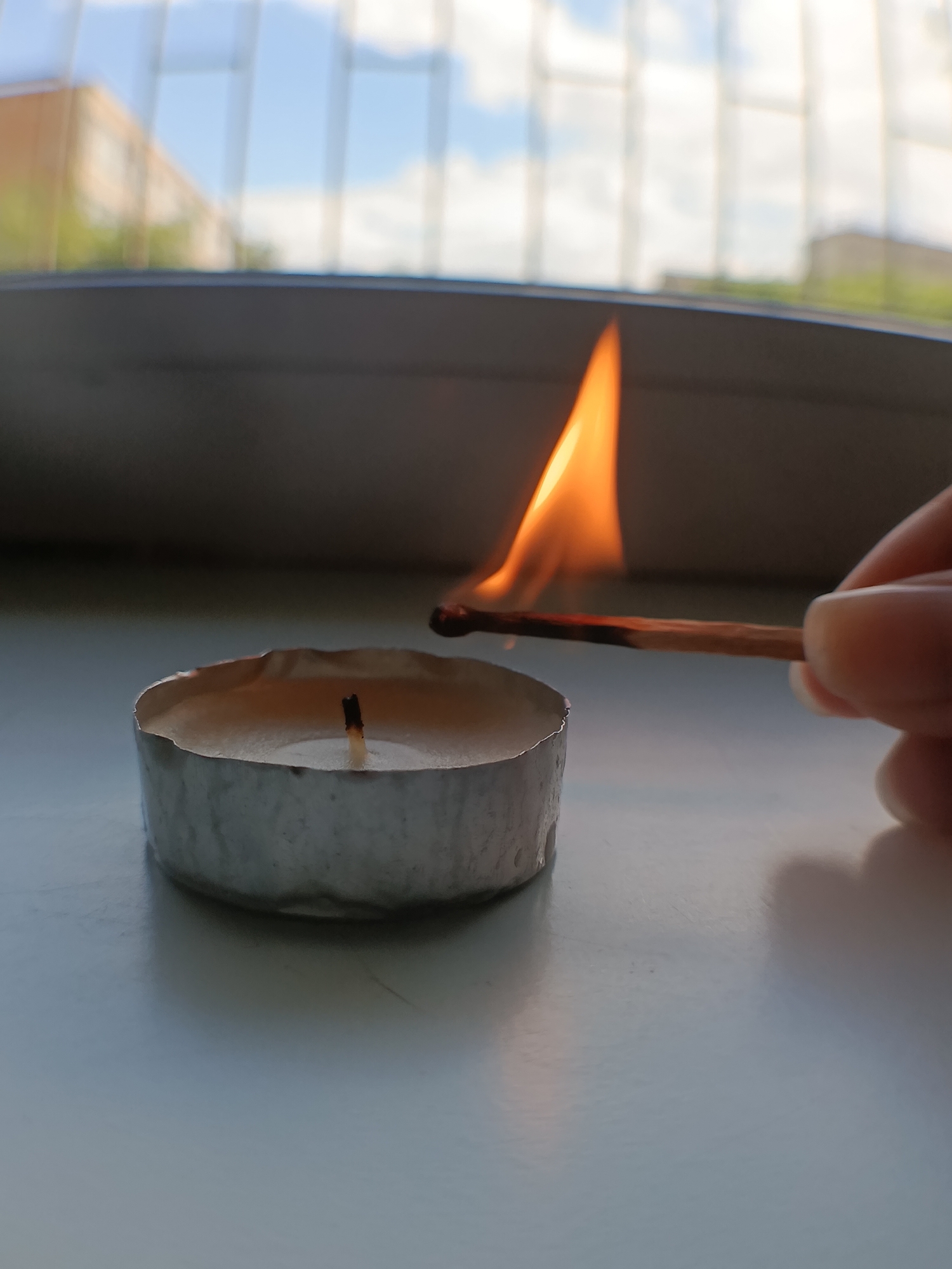 In the foreground, a tea light candle, in a silver foil holder, is being lit by a match. The match head is glowing red, and a flame is emerging from it, touching the candle's wick. A person's hand, holding the match, is visible on the right side of the image. The candle and match are placed on a light-colored, reflective surface. In the background, a blurry window shows the sky with clouds and the muted colors of buildings and trees, suggesting an urban or suburban setting.