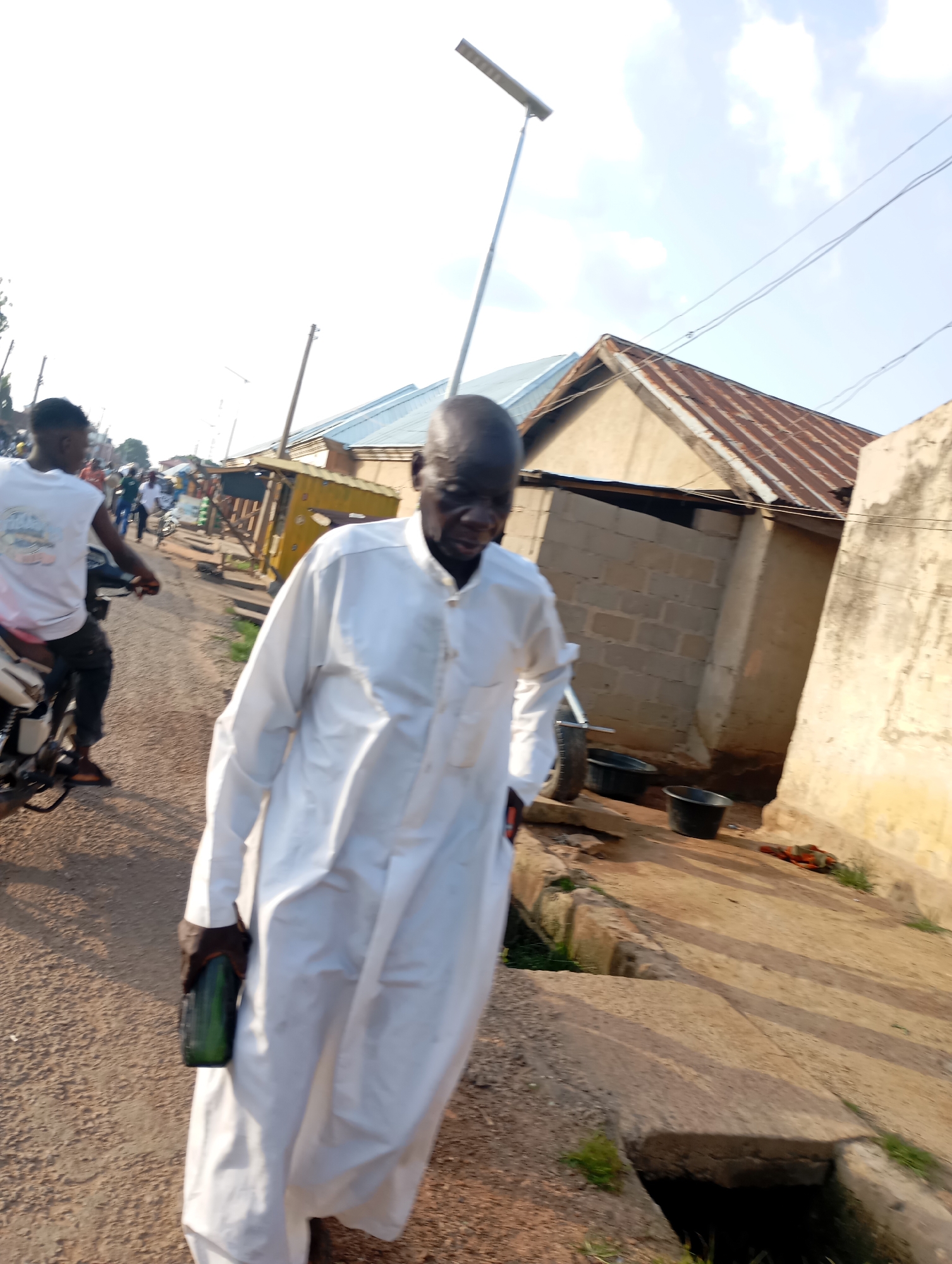 In Shendam, Nigeria, an older Black man wearing a flowing white robe walks down a dirt road. He is holding a dark green cylindrical object, possibly a bottle or container, in his right hand. To his left, a younger Black man on a motorcycle faces away from the camera, wearing a white tank top and dark pants. The background shows several buildings with corrugated metal roofs and concrete block walls. There are power lines overhead and what appears to be a solar-powered street light on a pole. The road is unpaved, and a concrete drainage channel runs along the right side of the road.