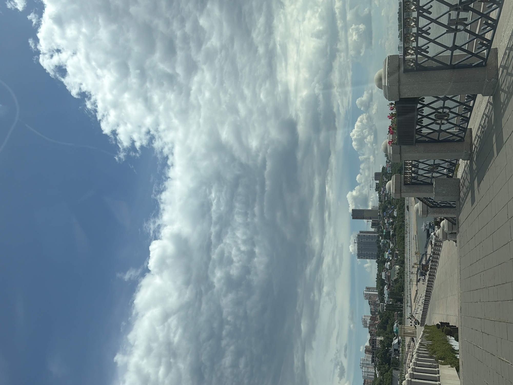 This media file depicts an outdoor scene in Ufa, Russia, likely taken from an elevated vantage point such as a balcony or promenade. The image is oriented vertically, showing a sweeping view of a cityscape and a vast sky filled with clouds.
The foreground features a stone railing with ornate metalwork, characteristic of a public walkway or scenic overlook. Planters with red flowers are visible along the railing, adding a touch of color. Beyond the railing, a paved pathway extends into the distance, bordered by greenery.
The middle ground reveals a cityscape with various buildings, including high-rise structures and smaller residential buildings, interspersed with trees. A body of water, possibly a river or lake, is visible, with a quay or embankment running alongside it. A few boats are docked.
The sky dominates a significant portion of the image, filled with dynamic cloud formations. Cumulus clouds, some appearing puffy and white, contrast with more elongated, textured clouds, suggesting varied atmospheric conditions. The presence of some haze or thinner cloud layers in the distance hints at the time of day, likely late morning or early afternoon, with the sun filtering through.
There are no people or specific activities clearly visible in the image. The overall mood is serene and expansive, emphasizing the grandeur of the natural sky and the urban landscape. The location cues, such as the architecture of the railing and the style of buildings, combined with the explicit mention of Ufa, Russia, provide a clear geographical context. No visible text is present in the image.