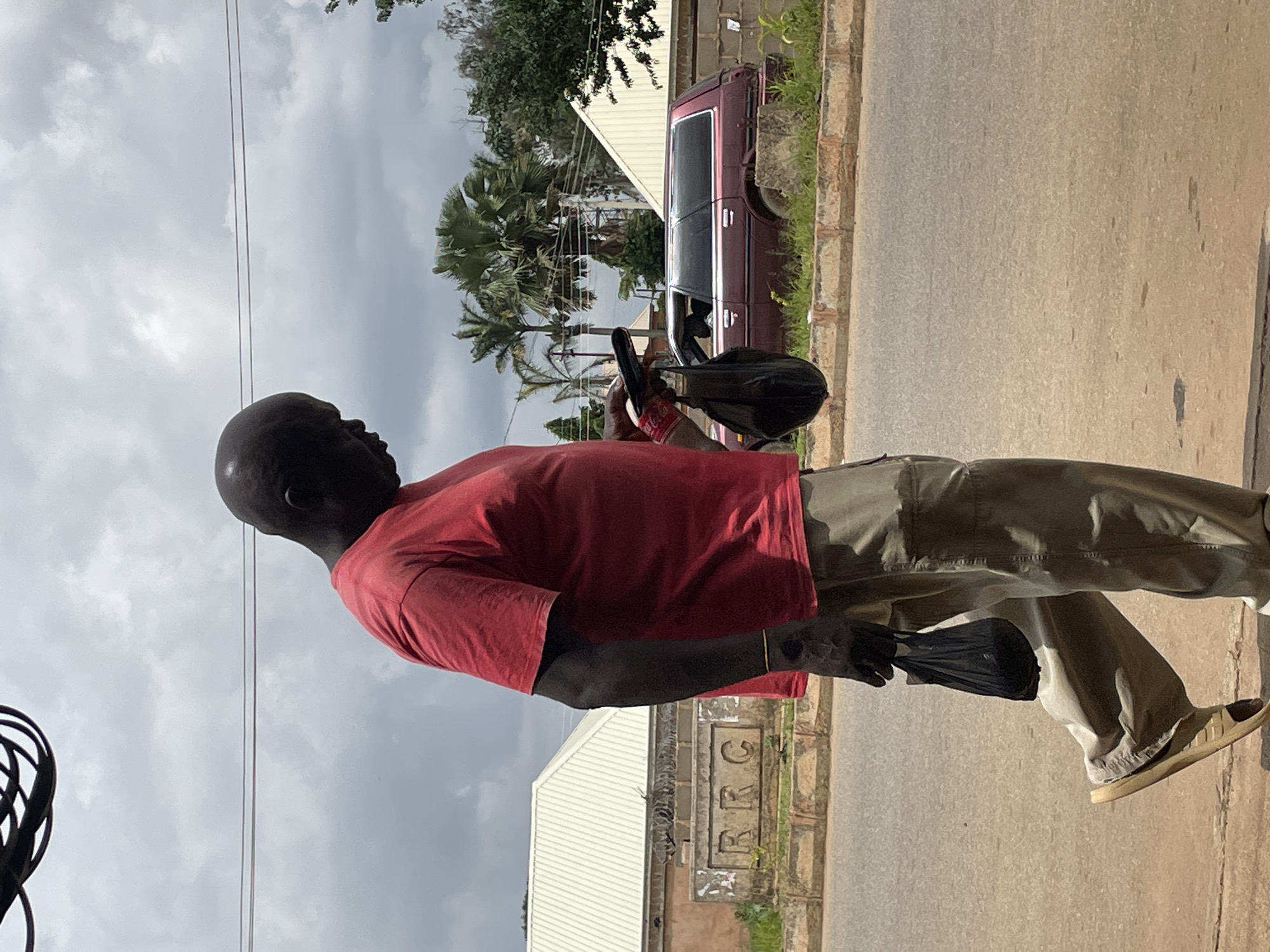 A bald Black man wearing a red t-shirt and tan cargo pants walks along a paved road in Bukuru, Nigeria. He carries two black plastic bags, one in each hand. The man is looking down at a mobile phone he holds in his right hand. A maroon station wagon is parked on the side of the road behind him, with palm trees and other vegetation visible in the background. A building with a white roof and a sign that reads "RRC" is to the left of the man. The sky is cloudy.