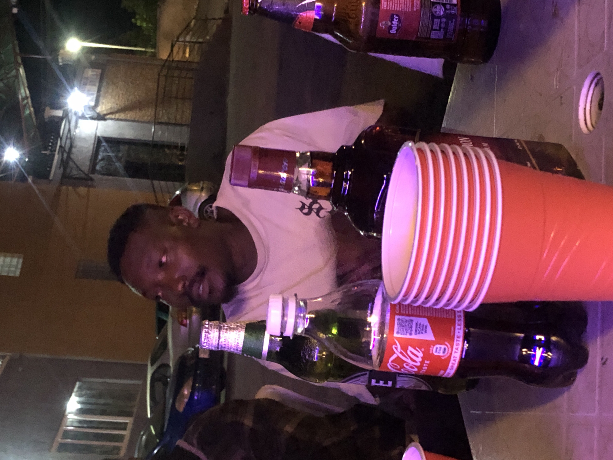 The image shows a man sitting at a table outdoors at night. He is wearing a white t-shirt and has a mustache. In front of him on the table are several drinks, including a bottle of Coca-Cola, a green bottle that appears to be a beer bottle, a bottle of what looks like whiskey, and a stack of red plastic cups. There is also a bottle of Gulder beer in the background. The environment appears to be a bar or outdoor gathering space, with cars and buildings visible in the background, illuminated by artificial lights. The lighting is dim and tinted purple in some areas.