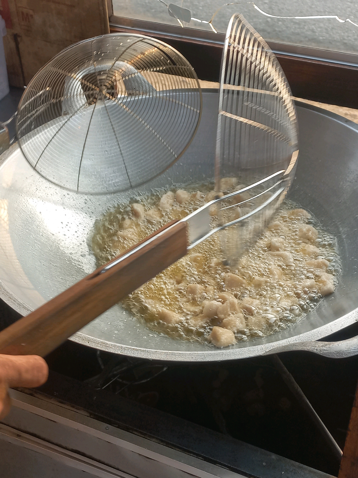 In Baturetno, Indonesia, a close-up, high-angle shot captures the process of deep-frying food. The scene takes place outdoors, possibly at a street food stall, indicated by the warm lighting and the presence of a window frame with cracked glass in the background. A large, metallic wok is filled with bubbling hot oil, in which numerous small, golden-brown pieces of food are being fried. A wooden-handled metal spider strainer, likely being used to turn or remove the food, is positioned above the wok. To the left, another metal strainer with a domed, mesh surface rests on the rim of the wok. The food appears to be in bite-sized pieces, suggesting it could be snacks or appetizers. The overall impression is one of a bustling food preparation environment.