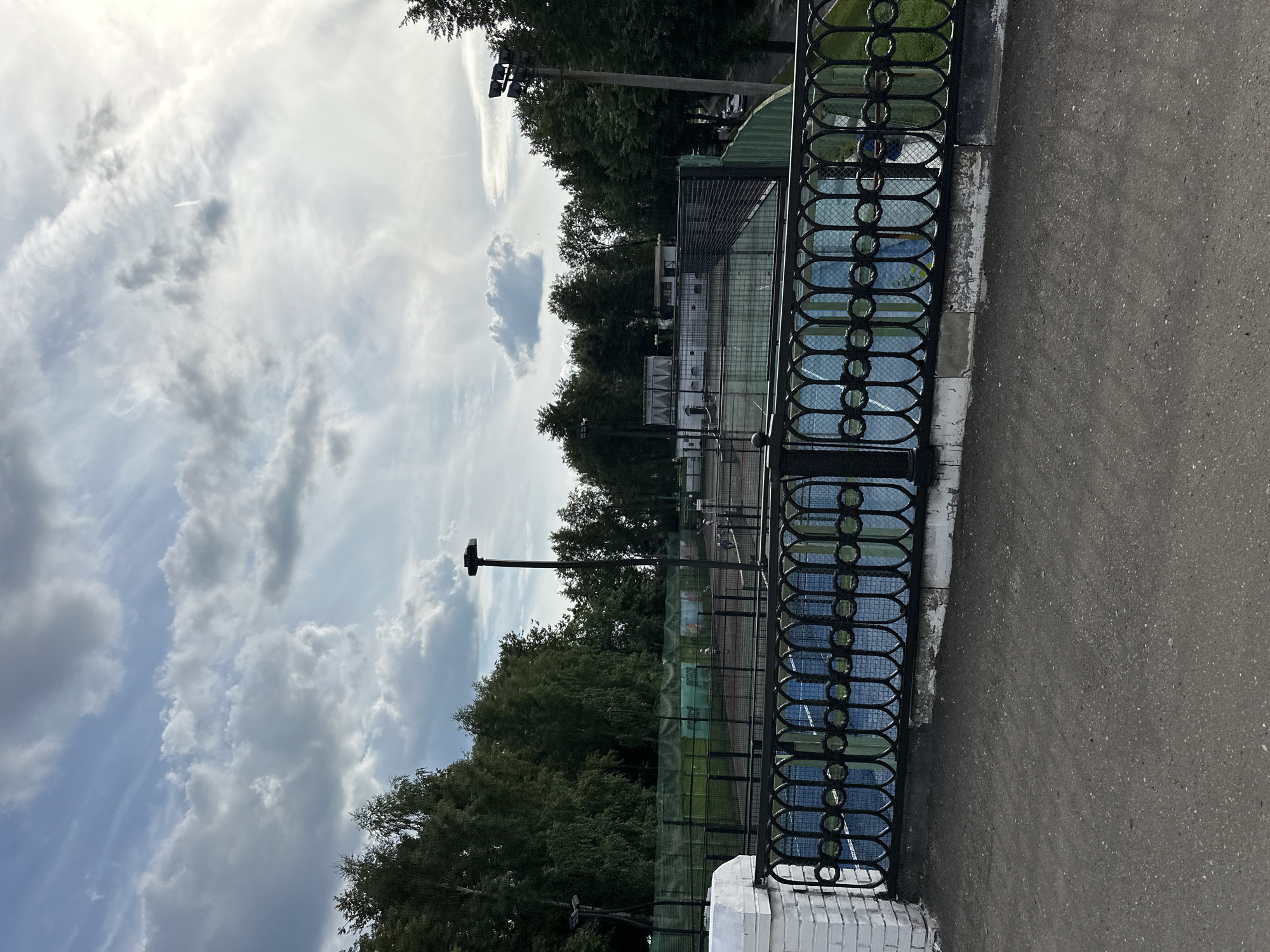 The image depicts an outdoor sports facility, likely a tennis or padel club, located in Zhukovo, Russia. In the foreground, a grey asphalt path leads to a decorative black metal fence with an oval link pattern, partially obscuring a view of several blue and green courts. Beyond the fence, the courts are enclosed by green netting. In the mid-ground, several people are visible on the courts, some appearing to be playing. A white building with arched windows is situated behind the courts. The background is dominated by a dense line of mature green trees. Above, the sky is partly cloudy with a mix of white and grey clouds, illuminated by sunlight suggesting it is daytime. Lighting poles with multiple lamps are present, indicating the facility may be used in the evening. A white brick structure, possibly a pillar, stands to the left of the fence.