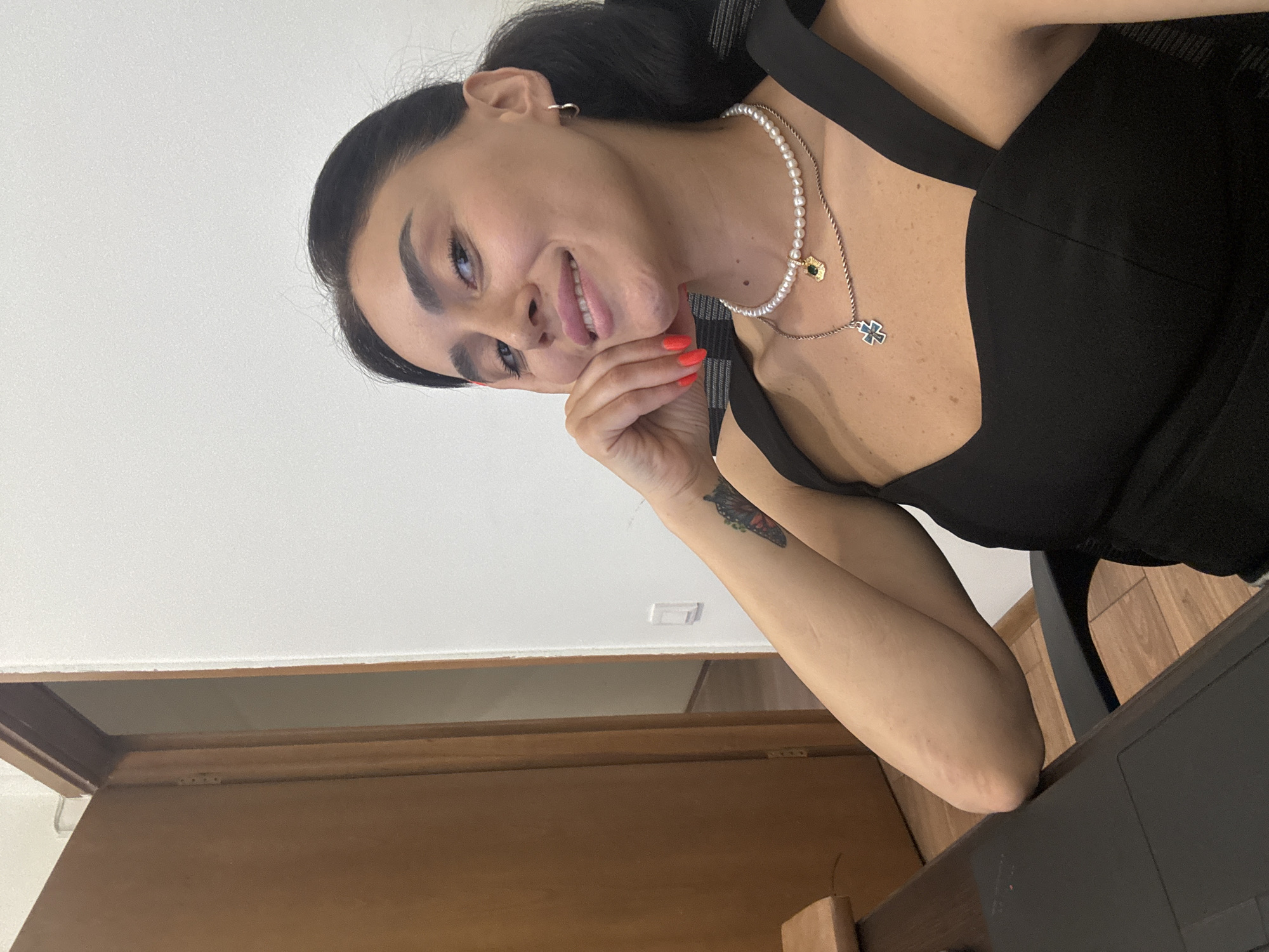 A woman is sitting at a desk with her hand resting on her chin, looking at the camera. She is wearing a black top and two necklaces. She has dark hair pulled back in a ponytail and is smiling. There is a tattoo of a butterfly on her left arm. The background shows a doorway and a white wall. The setting appears to be an office or indoor space. There is no indication that this image was taken in Tbilisi, Georgia.