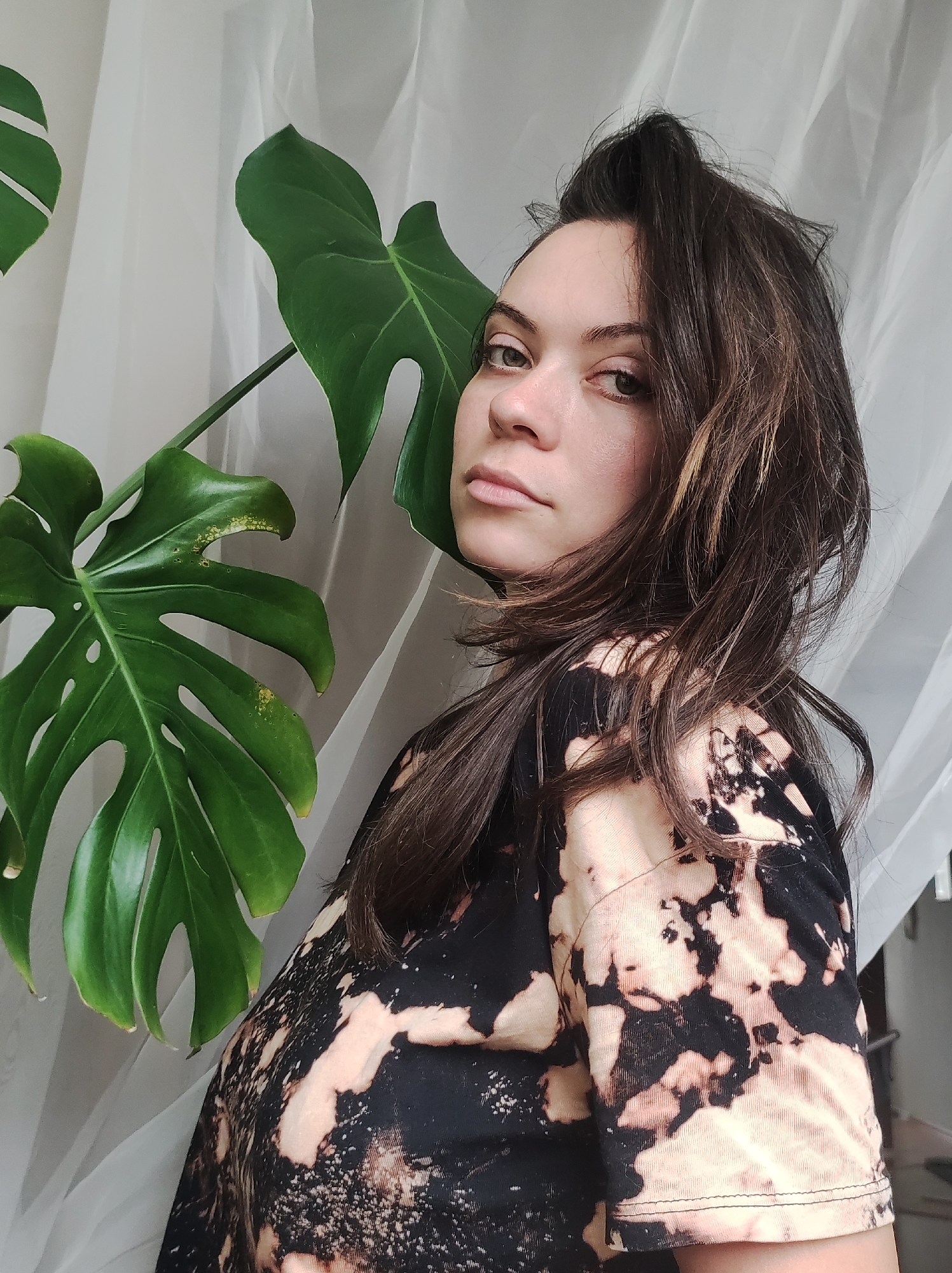 A medium shot captures a woman posing next to a large monstera plant against a backdrop of sheer white curtains. The woman has fair skin, dark brown hair with lighter highlights, and is wearing a t-shirt with a bleached tie-dye pattern in black and beige. The monstera plant has several large, vibrant green leaves with characteristic fenestrations. The setting appears to be indoors, with natural light filtering through the curtains. The specific location within the city of Chaikovsky, Russia cannot be determined from the image alone.