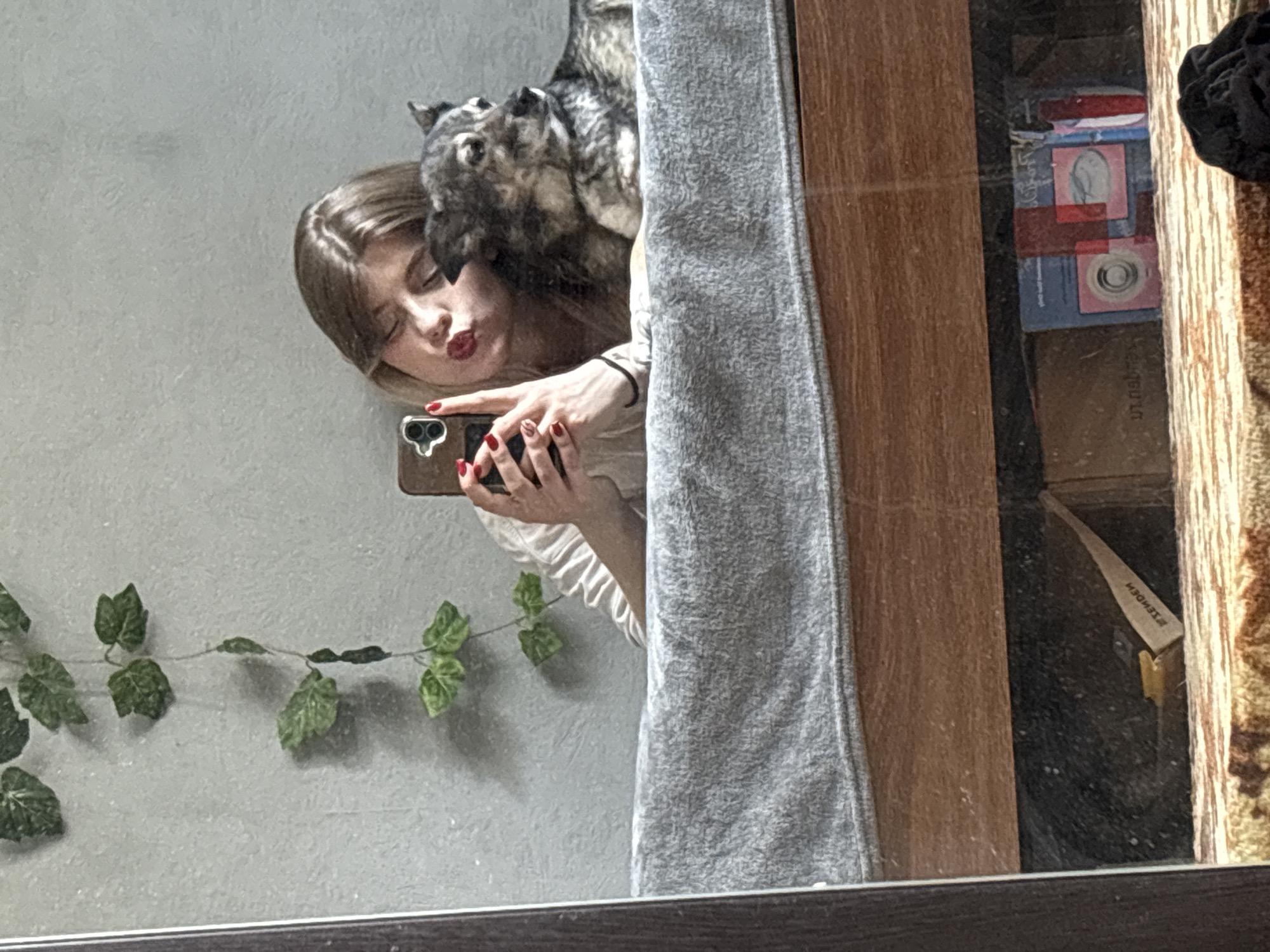 A young woman with blonde hair and red nail polish is taking a mirror selfie with her dog. She is leaning on her dog, who is lying down next to her. The woman is wearing a white shirt, and her lips are puckered. The dog is a medium-sized breed with gray and black fur. They are both on a bed, covered with a gray blanket. In the background, there is a gray wall with a string of artificial green leaves hanging from it. The reflection in the mirror also shows the wooden frame of the mirror and the underside of the bed, which contains some scattered items.