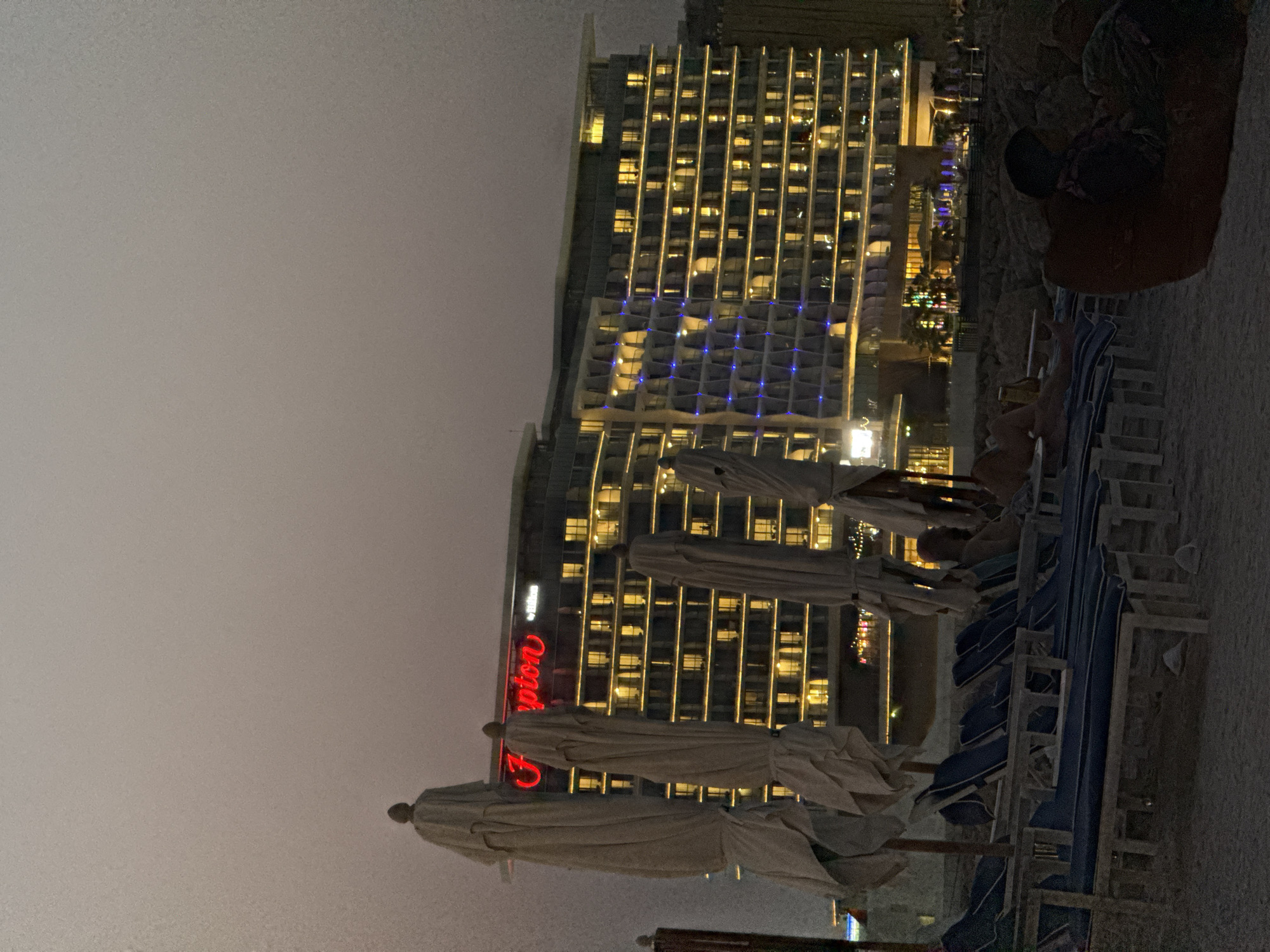 The image shows a twilight scene on a beach in Ras Al-Khaimah, United Arab Emirates. A large, modern hotel with illuminated windows dominates the background, its "Hampton" sign prominently displayed in red neon. In the foreground, several beach loungers with blue cushions are arranged on the sand, some covered with beige beach umbrellas. A few individuals are visible on the loungers, with one person partially visible in a beanbag chair in the lower right corner. The sky is a muted gray, suggesting the transition from day to night. The environment appears to be a resort or hotel beach area.