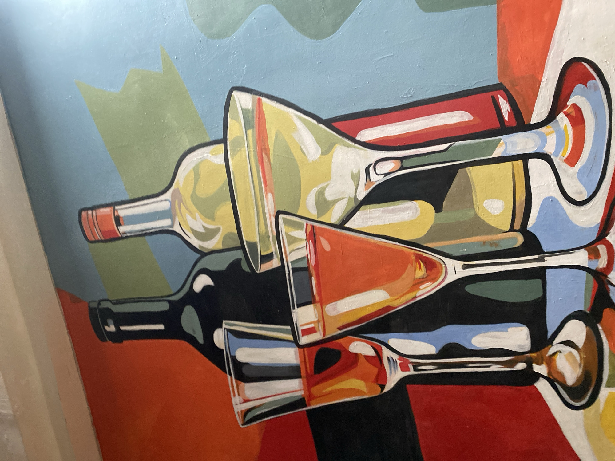 This is a close-up, eye-level, medium shot of a painting featuring two bottles and three glasses of drinks. The painting has a stylized, flat, graphic art aesthetic with bold outlines and bright, contrasting colors.
In the background, two bottles are depicted. The bottle on the left is dark, possibly black or very dark blue, with a slender neck and a cap. The bottle on the right is lighter, appearing to be a pale yellow or green, with a red cap. Behind the bottles, abstract shapes in shades of orange, green, and blue create a dynamic backdrop.
In the foreground, three glasses are prominently displayed. Two of the glasses are on the left and are of similar shape, with tall stems and conical bowls. They contain amber-colored liquids. The third glass, on the right, is a martini glass with a wide, shallow bowl and a long stem, and it contains a pale yellow liquid with a reddish-orange stripe across the top. The glasses are rendered with bright highlights and bold, colored lines that suggest reflections and the liquid's contents.
The overall impression is a vibrant and abstract still life of alcoholic beverages. There is no indication of a specific location within the city of Howayi, Nigeria. The painting appears to be a decorative piece, perhaps a mural or artwork on a wall.