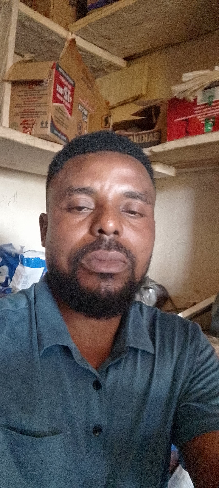 In a dimly lit room, a man with dark skin, a short haircut, and a beard faces the camera. He wears a blue collared shirt with two dark buttons visible. The background is cluttered with various cardboard boxes stacked on shelves, suggesting a storage or warehouse environment. The lighting is dim, casting shadows and contributing to a slightly grainy quality of the image. The overall setting appears to be indoors. The image location is unknown.