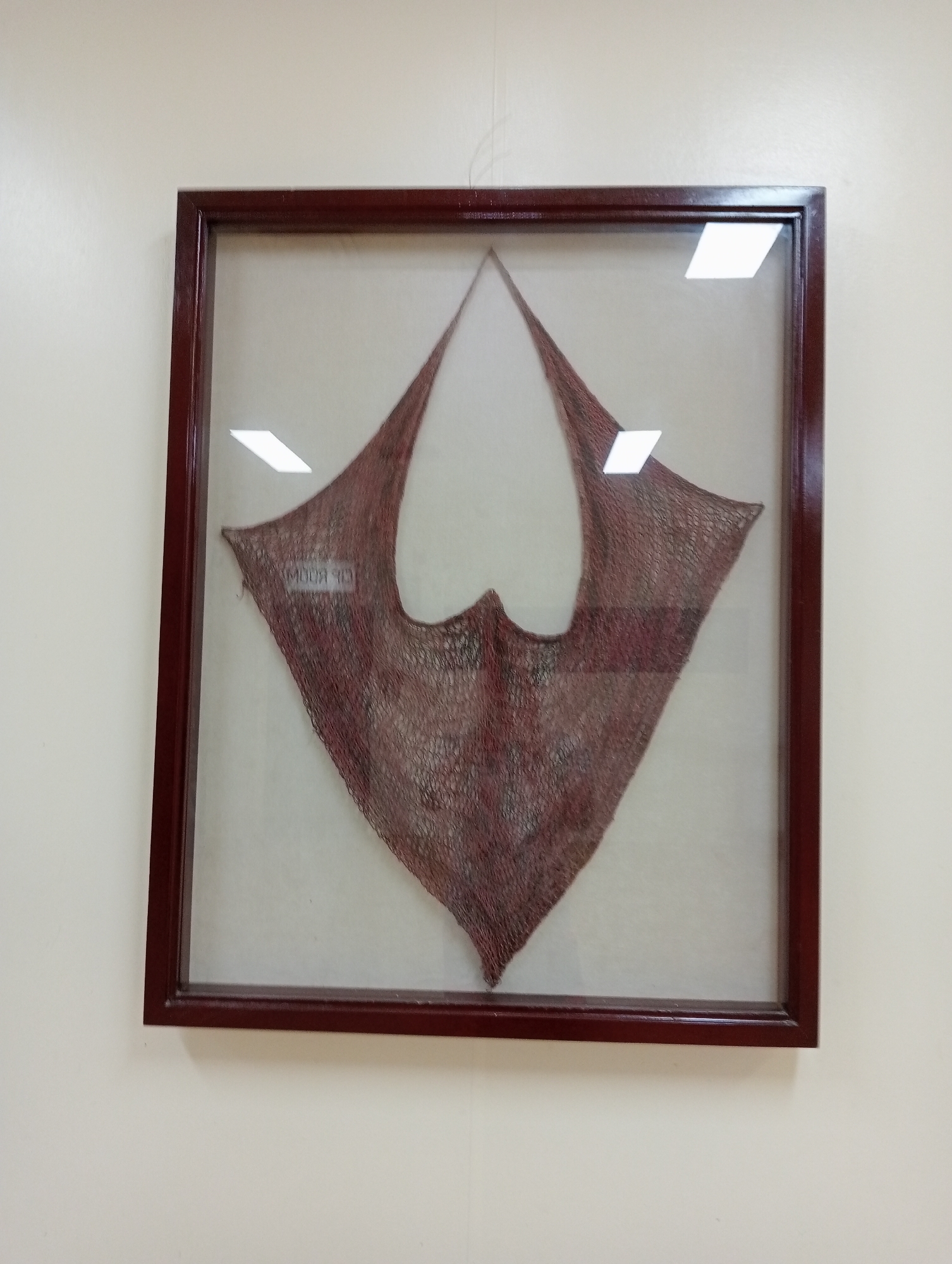 An antique, maroon-colored, framed textile is displayed on a white wall. The textile, likely a shawl or a similar garment, has a maroon and red patterned weave with a net-like texture. It is shaped like a triangle with a curved bottom edge, and the top point is folded. There is a label visible on the textile that reads "MODA 313". The frame is made of dark wood with a polished finish. The lighting creates several bright reflections on the glass of the frame.