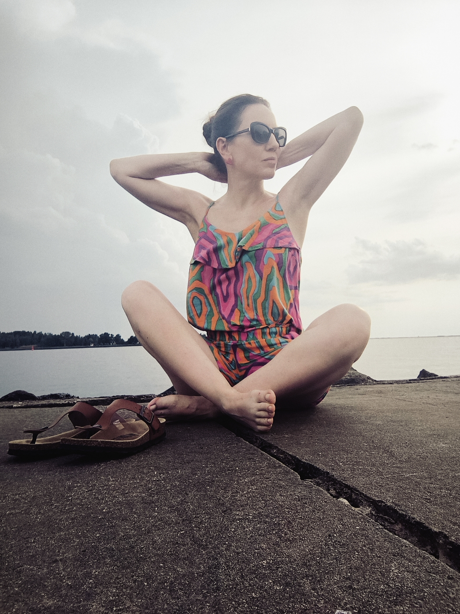 A woman is sitting cross-legged on a concrete surface near a body of water, with trees and buildings visible in the distance. The sky is overcast. She is wearing sunglasses and a colorful patterned romper, and her hands are behind her head. A pair of brown sandals rests on the concrete to her left.