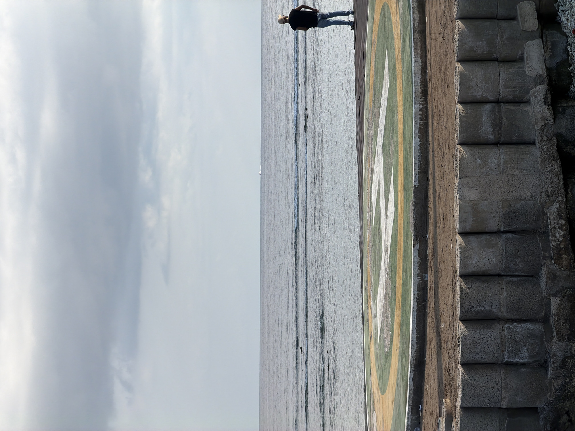 A full shot captures a helipad on a waterfront with a person standing on it looking out at the ocean. The helipad is circular with white, yellow, and green markings and is surrounded by a rough, sandy area with concrete barriers. The ocean stretches to the horizon, with gentle waves and a small boat visible in the distance. The sky is overcast with a pale, cloudy atmosphere. The person is standing on the edge of the helipad, facing away from the camera, wearing a dark t-shirt, blue jeans, and a baseball cap. The location is identified as Unknown, Indonesia.