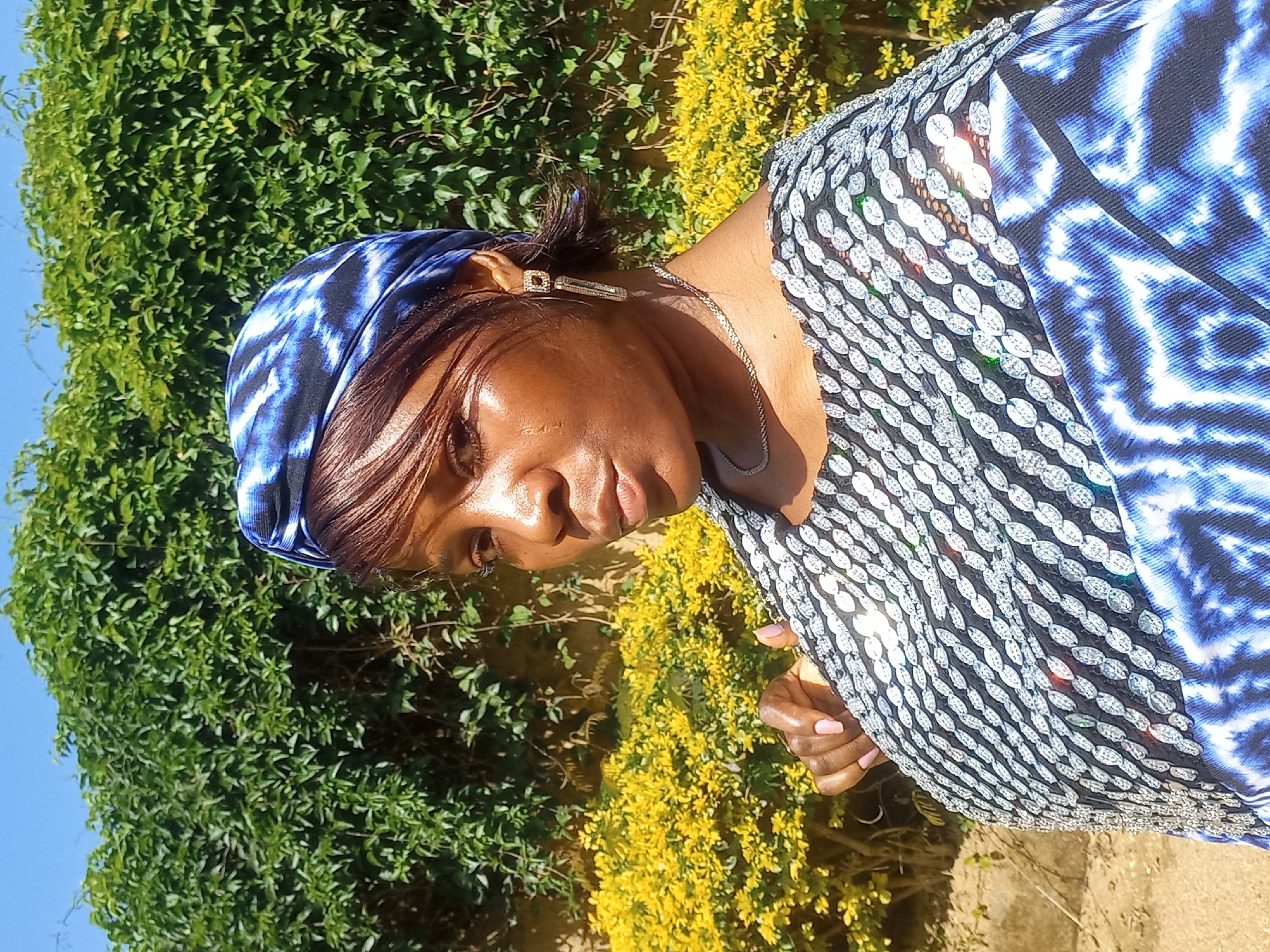 An outdoor, medium shot captures a woman posing for a selfie in what appears to be a garden setting in Jos, Nigeria. The woman is wearing a blue and white tie-dye headwrap and a black top adorned with reflective silver embellishments. She has light brown hair and is looking towards the camera with a neutral expression. Her left hand is visible, resting on her chest. The background consists of lush green foliage and a vibrant yellow flowering bush. The sky above is clear blue.