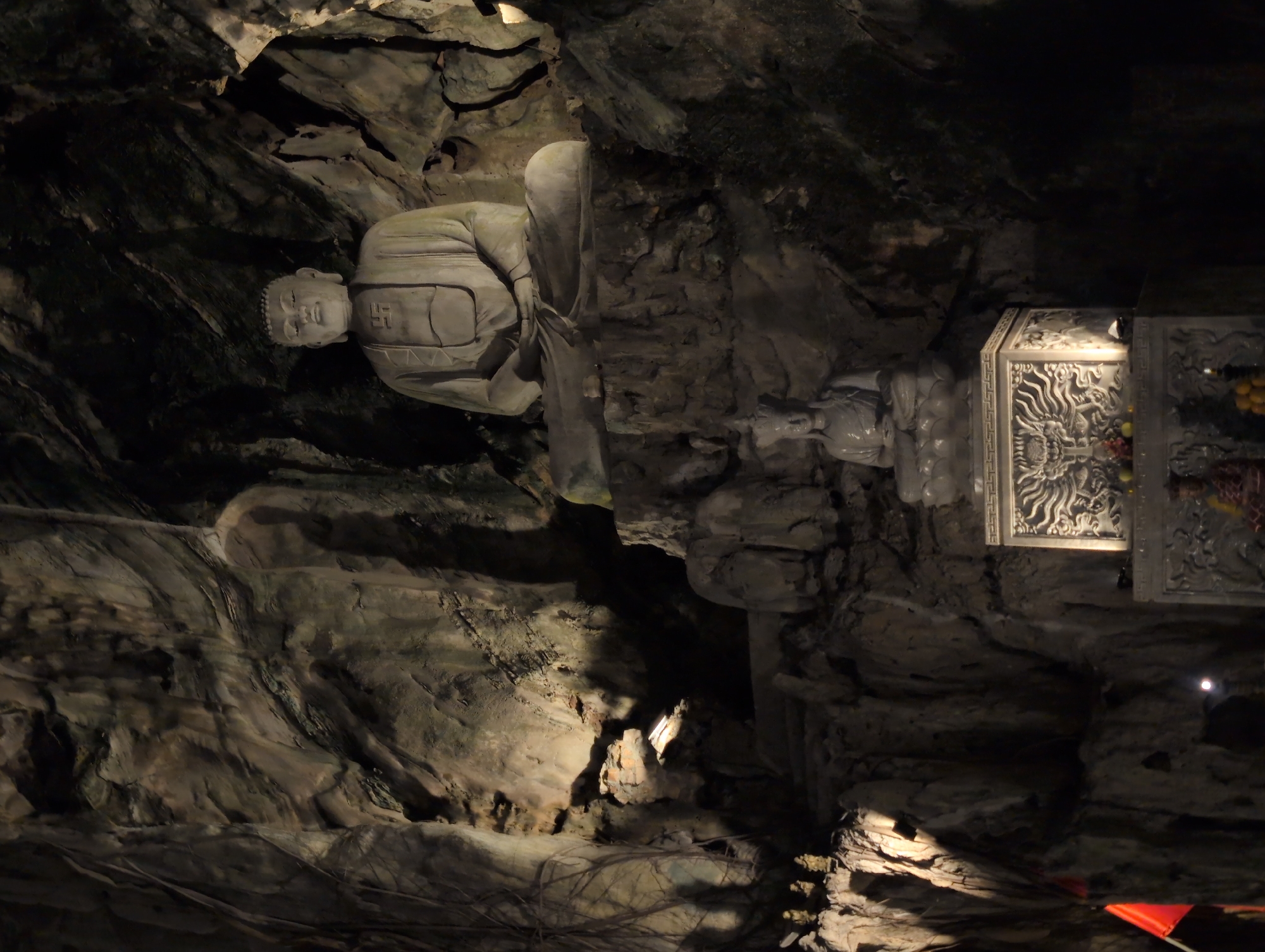 The image depicts a cave interior in the Marble Mountains, a popular tourist destination in the city of Đà Nẵng, Vietnam. The primary subject is a large, light-colored stone Buddha statue seated in a meditative pose within the cave. The statue is positioned on a rocky platform and is illuminated by artificial lighting that highlights its features and the cave's natural rock formations. Below the large Buddha statue, a smaller, also light-colored statue of a seated figure, possibly another Buddha or deity, is visible. In the foreground, a decorative stone altar or pedestal is present, featuring intricate carvings of a dragon. Fruits, likely offerings, are placed on and around the altar. The cave walls are dark and rough, with visible textures and shadows, suggesting a natural, uncarved environment. The overall atmosphere is solemn and spiritual.