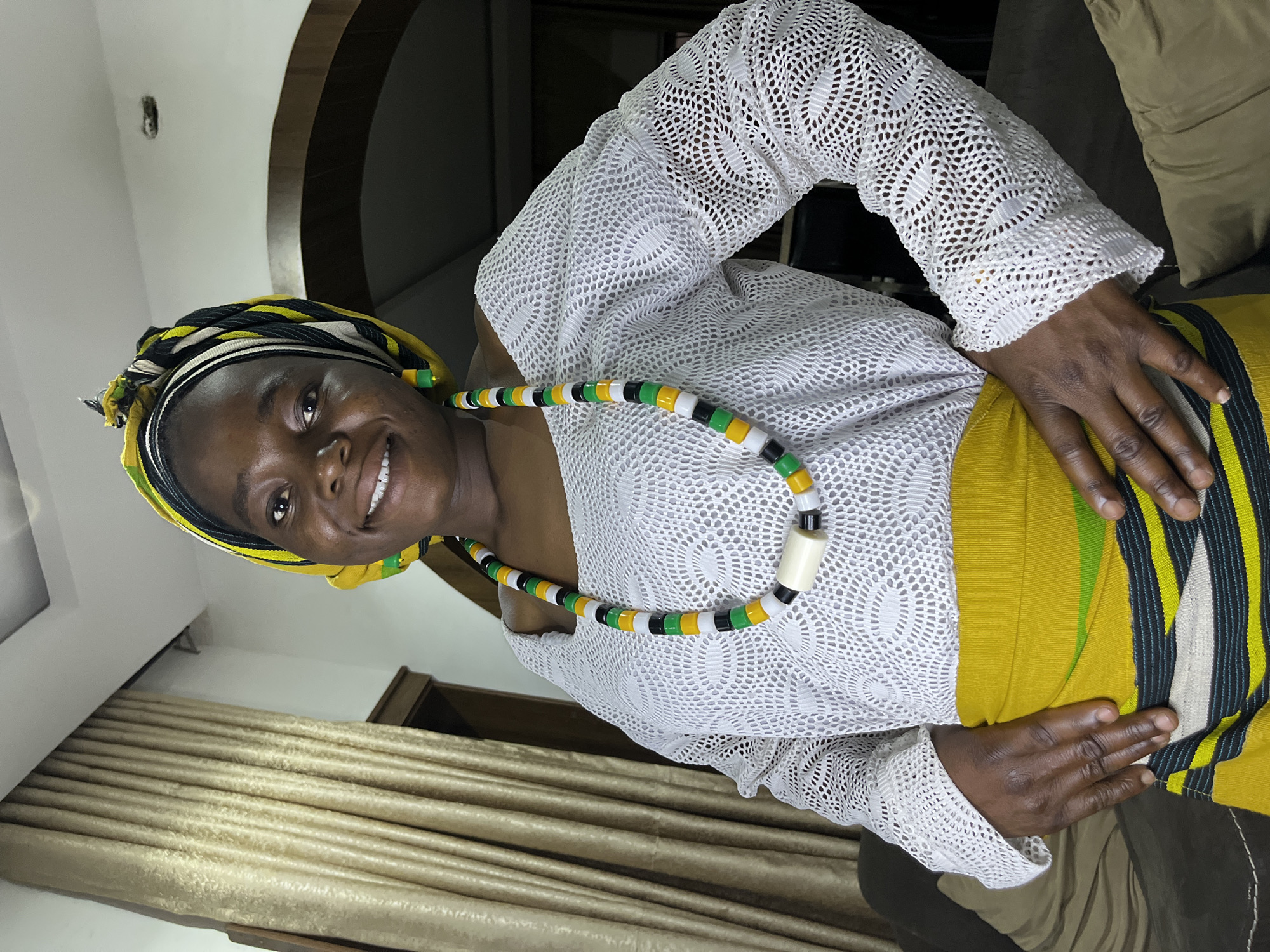 A woman is posing indoors. She is wearing a white, long-sleeved, lace top. She is also wearing a yellow, green, black, and white skirt. A necklace with yellow, green, black, white beads is around her neck. She is wearing a headwrap that is black, yellow, and green. Her hands are on her waist. Brown curtains are behind her. The location is presumably Jos, Nigeria.