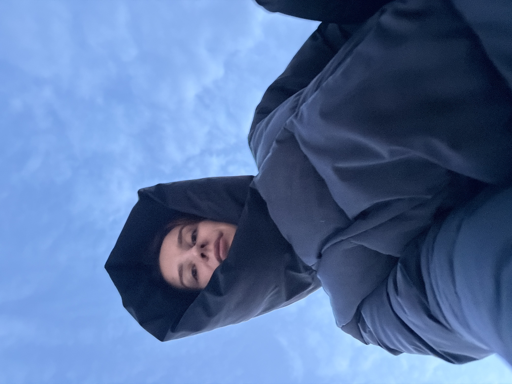 A low-angle, upward-facing shot shows a person's face framed by the dark hood of a puffy jacket against a pale blue sky. The jacket fills the majority of the frame. The person's expression is neutral. The location cannot be confirmed as the city of Chaikovsky, Russia, due to the lack of identifying features in the image.