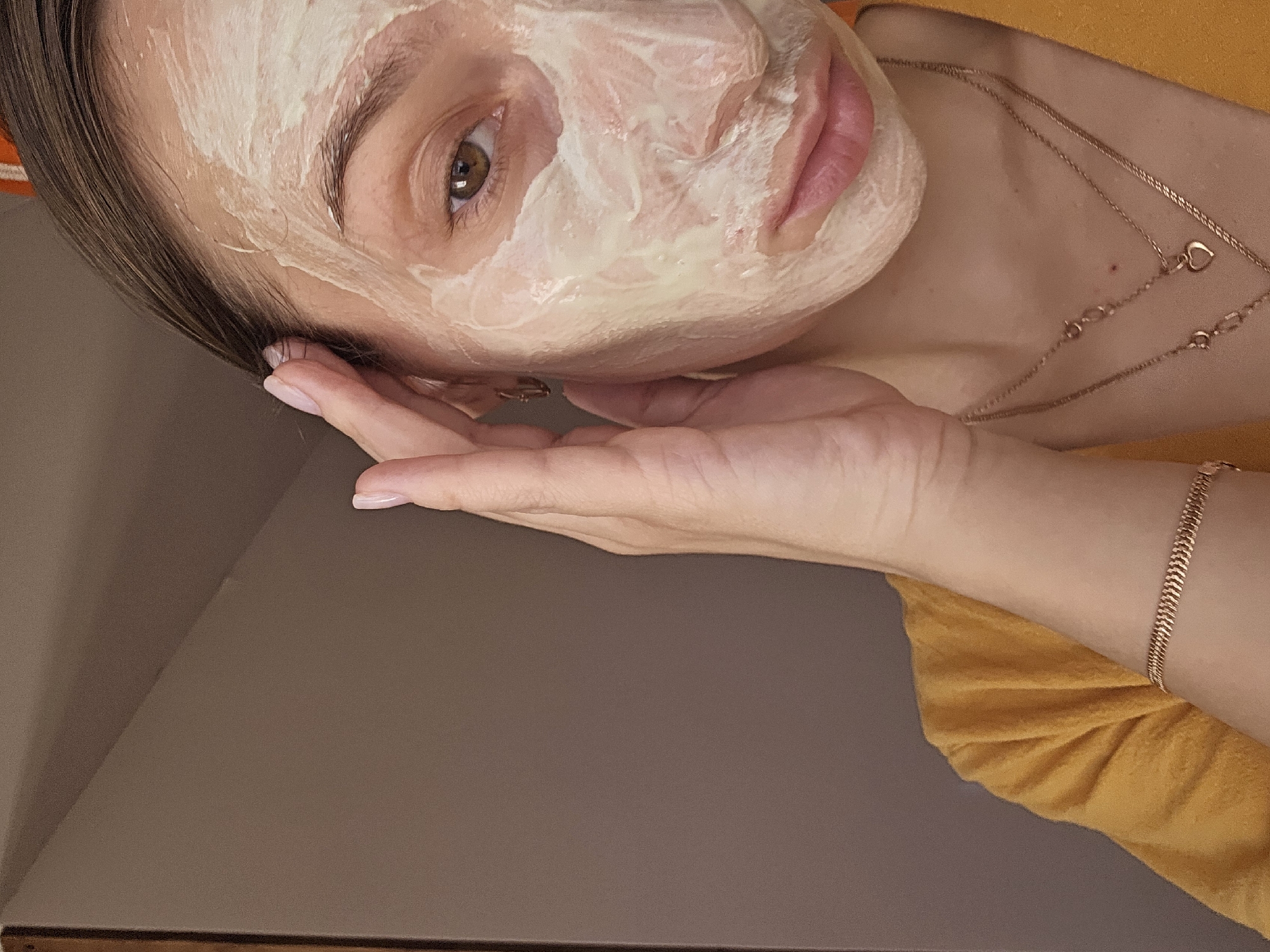 This image depicts the upper right quadrant of a female subject's face and upper torso in an indoor setting within the city of Chaikovsky, Russia. The subject's face is partially covered with a thick, light cream-colored facial mask, applied from the forehead down to the chin on the visible right side. Her right eye, with a brown iris, is open and looking towards the viewer. Her eyebrows are visible and appear groomed. Her lips are a natural pink color. Her dark brown hair is slicked back from her forehead, with a few strands near the hairline.
The subject's right hand, with manicured light pink nails, is raised, with fingers touching the temple and hairline area on the right side of her head. A small gold-colored hoop earring is present on her right earlobe. She wears a gold-colored, layered necklace with at least two chains, one featuring a small heart-shaped pendant. A gold-colored bracelet with a herringbone pattern is visible on her left wrist. She is wearing a yellow garment, possibly a shirt or robe, visible on her left shoulder and chest. The background consists of a plain, solid brown surface, possibly a wall or cabinet, with a darker vertical edge on the far left.