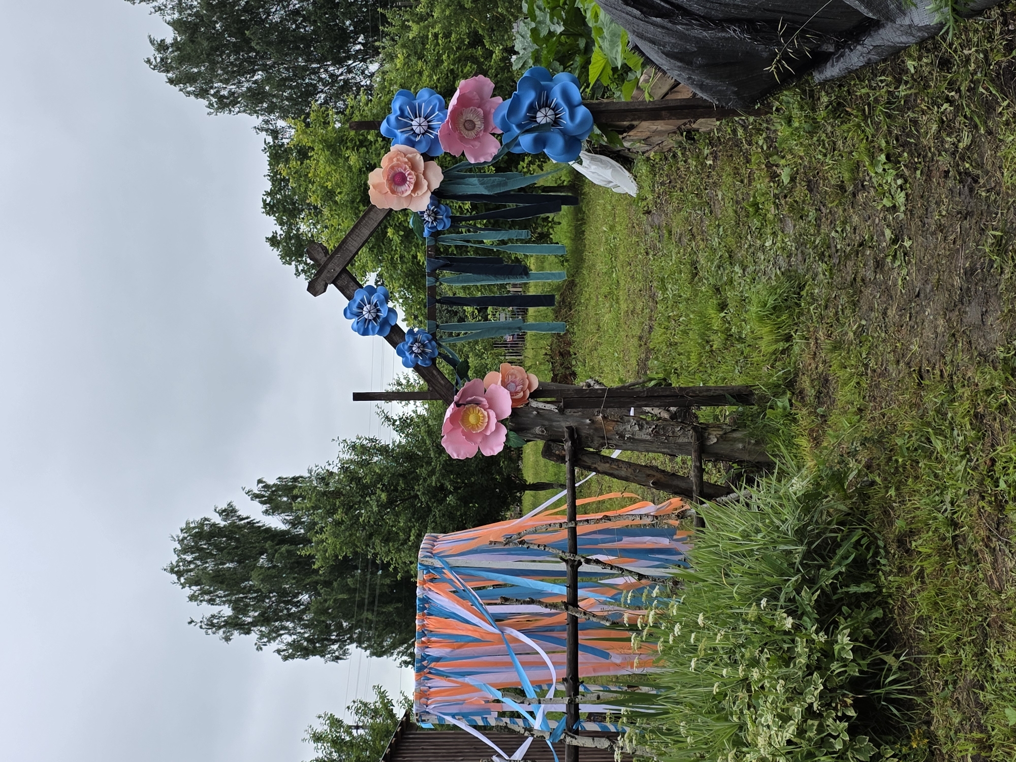 The image depicts an outdoor scene with a rustic wooden archway decorated with large, colorful paper flowers in shades of pink, peach, and blue. Strands of orange, white, and blue ribbons hang from a frame to the left of the archway, creating a decorative curtain. The ground is covered in green grass and appears damp. Lush green trees and foliage surround the area. In the background, a wooden structure resembling a shed or fence can be seen. The sky is overcast, suggesting a cloudy day. The scene appears to be set in a rural or garden environment, possibly in the village of Старые Быги, Russia. There are no people visible in the image.