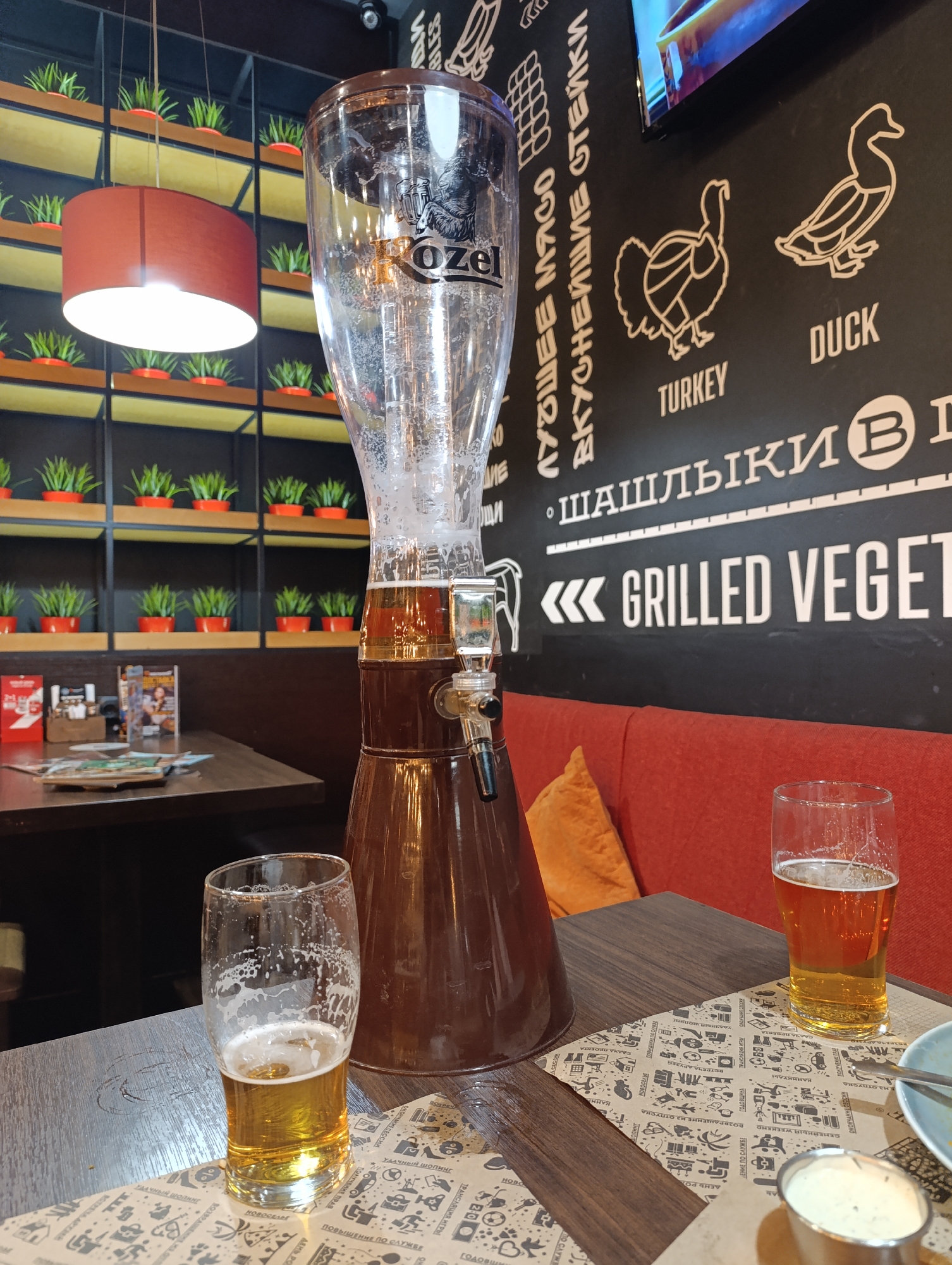 The image shows a restaurant interior, with a focus on a tall beer dispenser and two partially consumed glasses of beer on a table covered with paper patterned with doodles. The beer dispenser has the brand name "Kozel" on it. Behind the table is a black wall with the text "ЛУЧШИЕ МЯСО ВКУСНЕЙШИЕ ОСТЕЙКИ" and illustrations of a turkey and a duck, labeled accordingly. The wall also features the text "• ШАШЛЫКИ (В) <<< GRILLED VEGET". To the left, a table with several menus is visible. On the wall behind this table is a shelf lined with small potted plants. A red cylindrical lamp hangs from the ceiling above the potted plants. An orange couch is to the right of the beer dispenser, and there is a plate with a fork on the table as well as a small dish filled with dip.