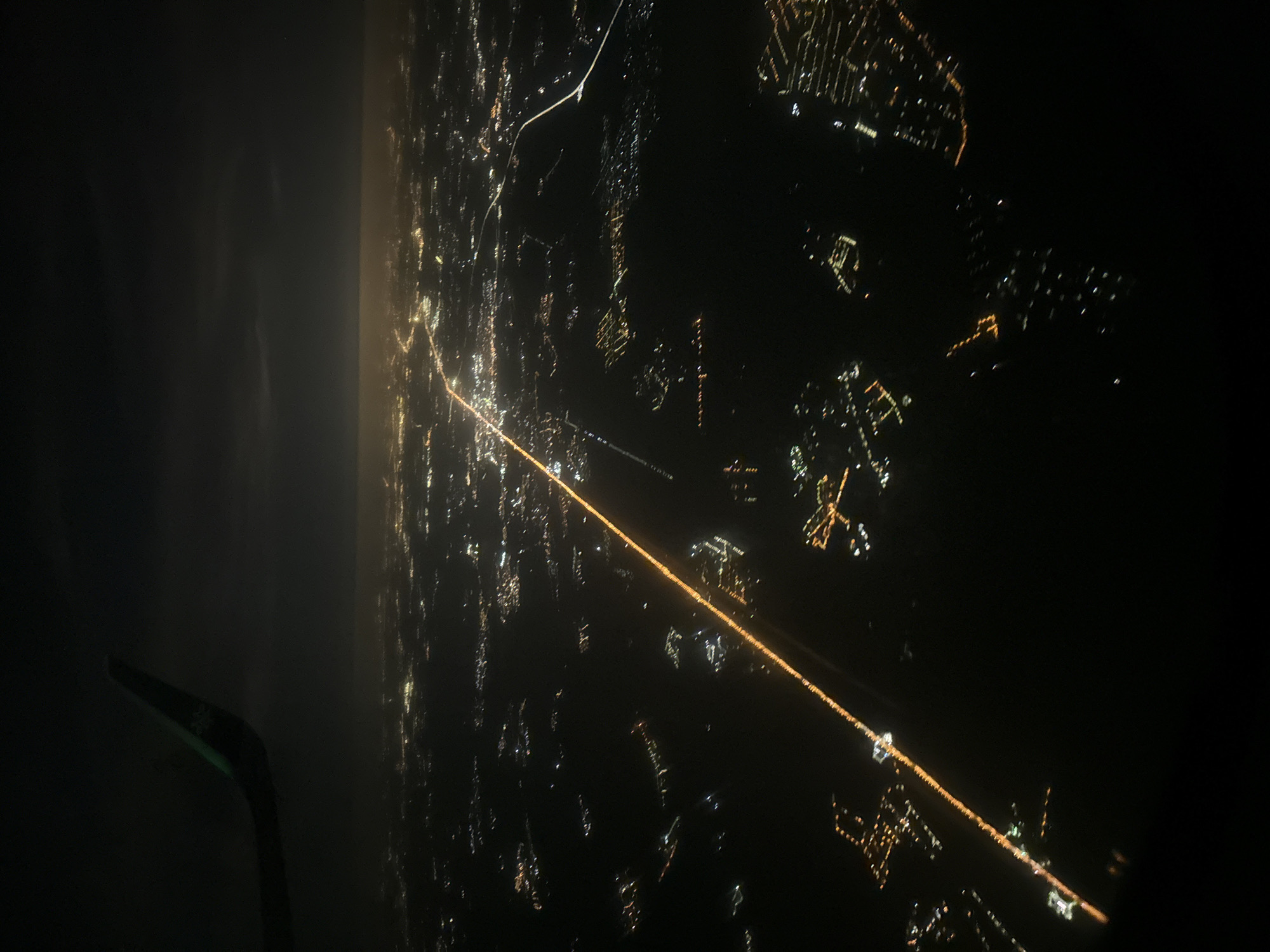 The image shows an aerial view of a city at night, likely taken from an airplane. The city is illuminated with numerous lights, forming a network of streets and buildings. A long, bright line of lights suggests a major road or highway. The wing of the plane is partially visible in the lower left corner. The location is not explicitly stated as Moscow, Russia, and cannot be confirmed based solely on the image.