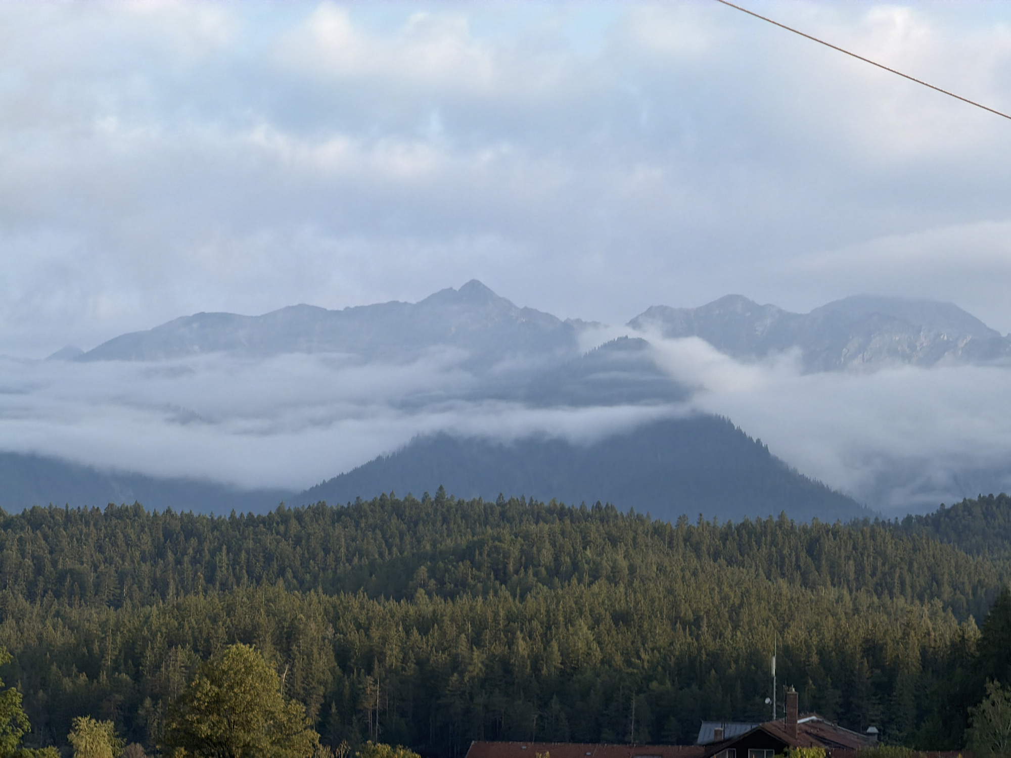 The image depicts a panoramic view of mountains and a dense forest in Grainau, Germany. In the foreground, a tiled roof of a building is visible, with a portion of a chimney and antenna. Behind the building, a vast expanse of green trees stretches across the middle ground, forming a thick forest that covers the slopes of smaller mountains. The background is dominated by a range of larger mountains partially obscured by mist and clouds, with hints of blue sky peeking through. The overall atmosphere is serene and natural, with the mist adding a sense of depth and mystery to the mountainous landscape.