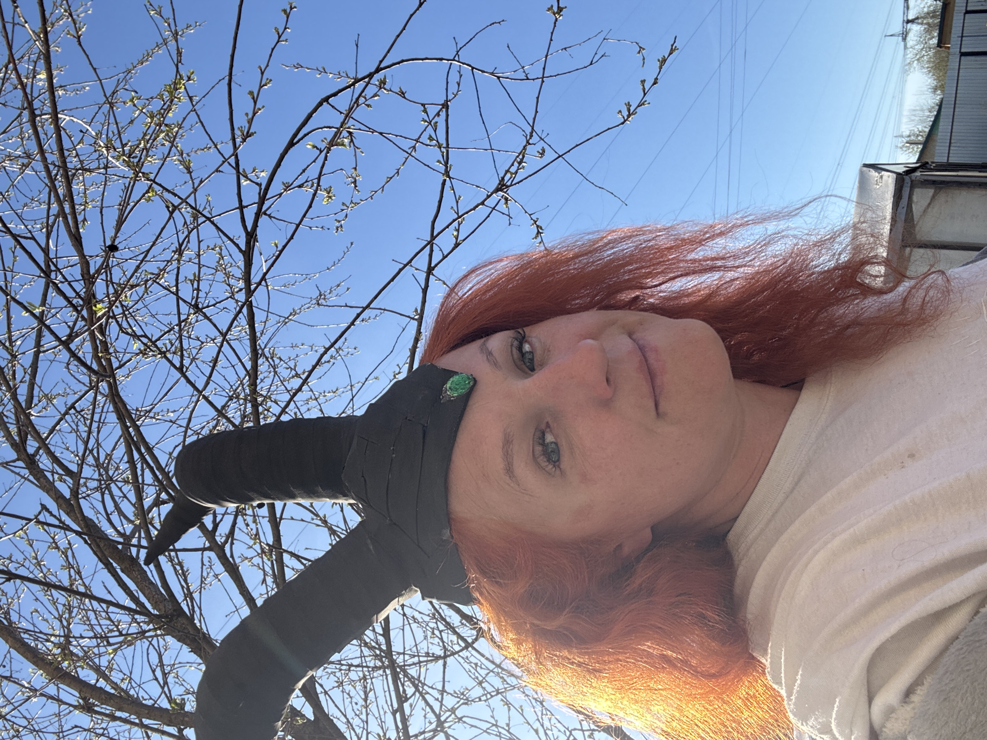 A woman with red hair and fair skin is wearing a black headdress adorned with two horns and a green stone. She is looking at the camera. The background features tree branches with budding leaves against a bright blue sky. A building and power lines are visible in the upper right corner. The image appears to be taken outdoors. The exact location in Chaikovsky, Russia, cannot be determined from the image alone.
