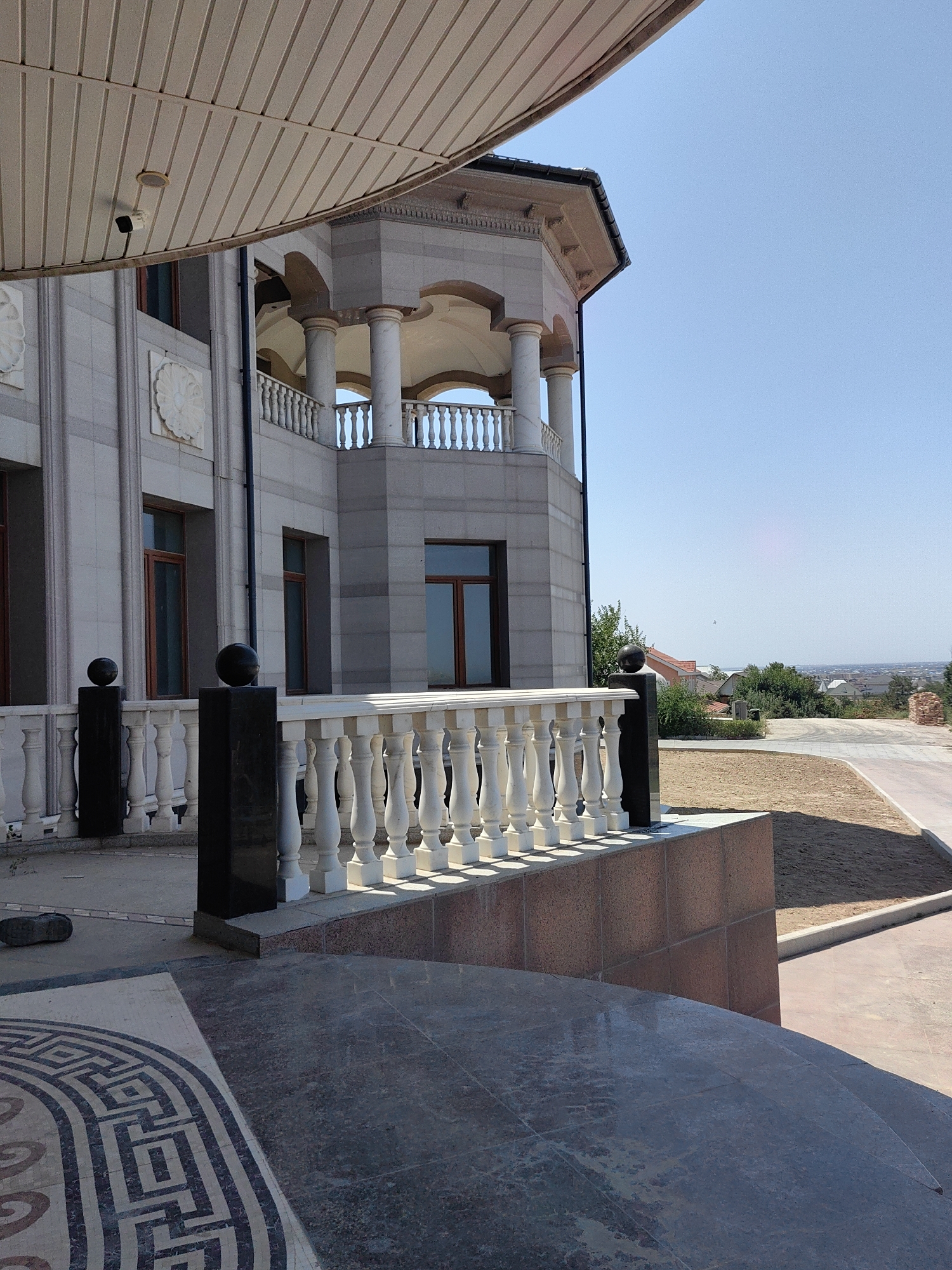 The image shows a view of a partially constructed or renovated building with classical architectural elements, including columns, arches, and a balustrade. The building is made of light-colored stone or concrete. In the foreground, there is a curved tiled surface and a mosaic pattern on the ground. A white balustrade with spherical finials adorns the edge of a raised area. Beyond the building, there is a street, some greenery, and houses in the background under a clear blue sky. There are no people visible in the image. The scene suggests a sunny day.