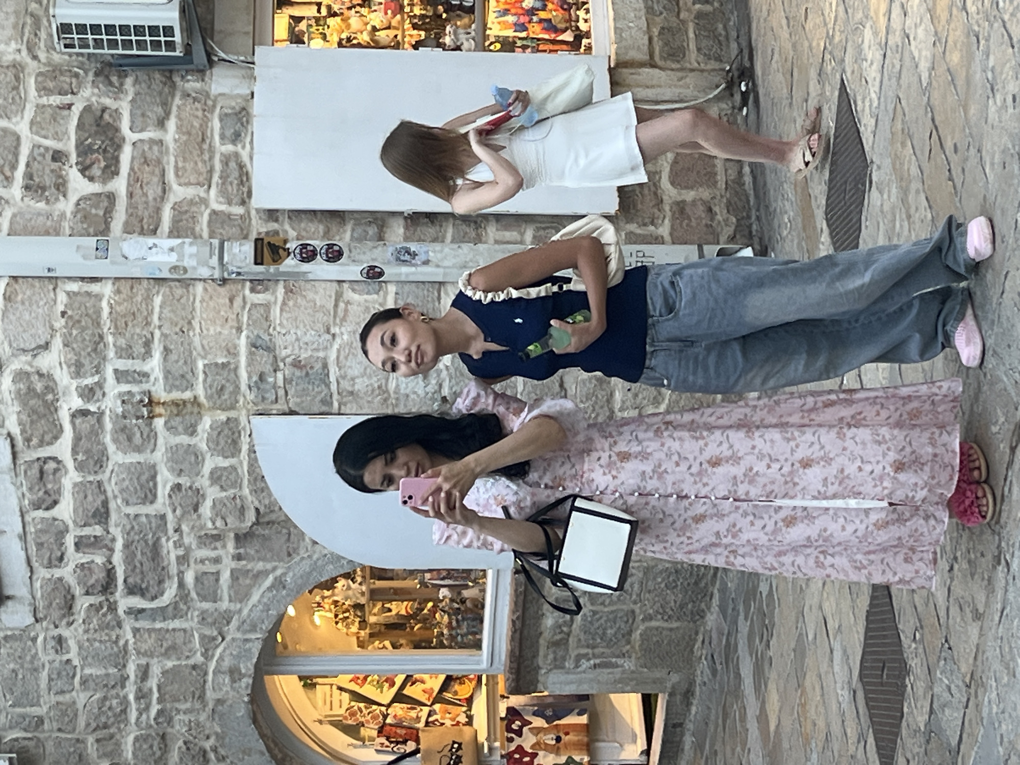 The image depicts a street scene in Budva, Montenegro, with three young women. The woman on the left, wearing a long pink floral dress and pink furry slides, is holding a pink smartphone and taking a picture. She has a black and white handbag with her. To her right, a woman in a navy blue sleeveless top and baggy jeans holds a green bottle and looks upwards. In the background, a third woman in a white dress and sandals is partially visible, holding a white tote bag and a blue bottle. The setting is a cobblestone street with stone buildings and shop windows displaying merchandise. An air conditioning unit is visible on the building to the right.