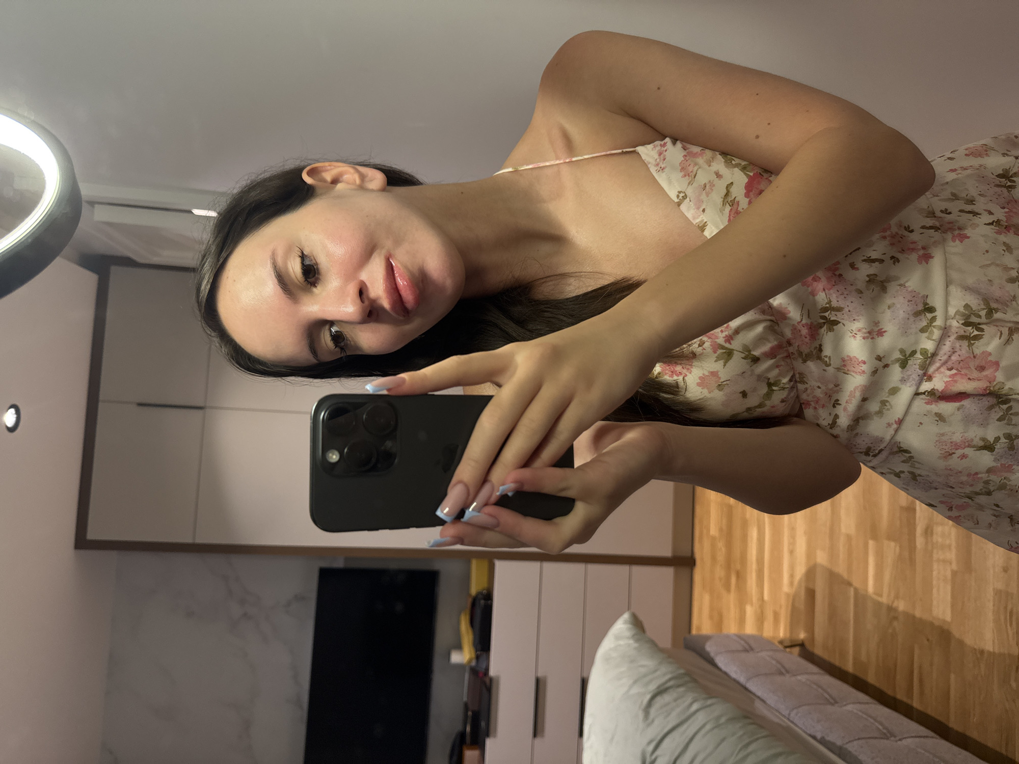A young woman is taking a selfie in a room with wood floors and a marble-patterned wall. She is wearing a floral dress and has light blue nail polish on her long, artificial nails. She is holding a black smartphone in her right hand, and her left hand is resting on the phone. A ring light is visible in the upper right corner of the image. The room appears to be a bedroom, with a bed on the left side of the frame. A dresser and a closet are visible in the background.